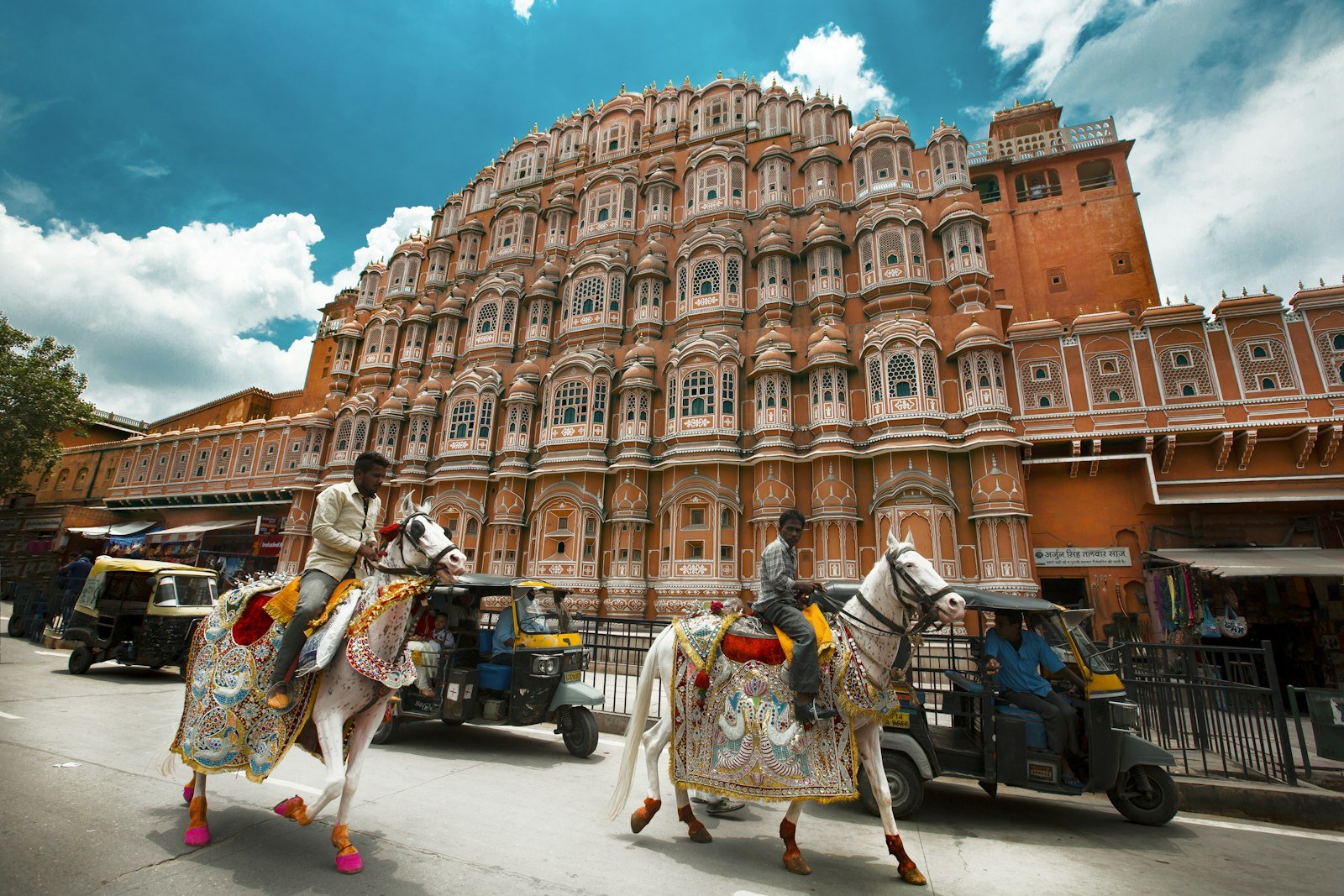 Hawa Mahal on Hawa Mahal Road in Jaipur with decorated horses and street traffic under a blue sky