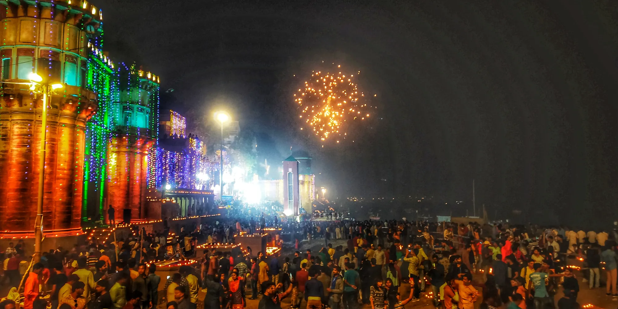 Crowds on a lamp-lit Varanasi ghat during Dev Deepawali with fireworks over the Ganga