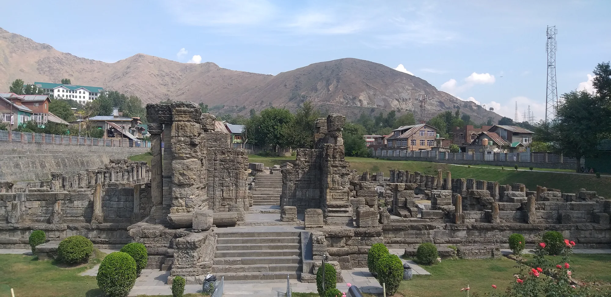 Stone ruins of the 9th-century Avantiswami temple at Avantipora, with steps up to the central shrine, a green lawn and brown hills behind