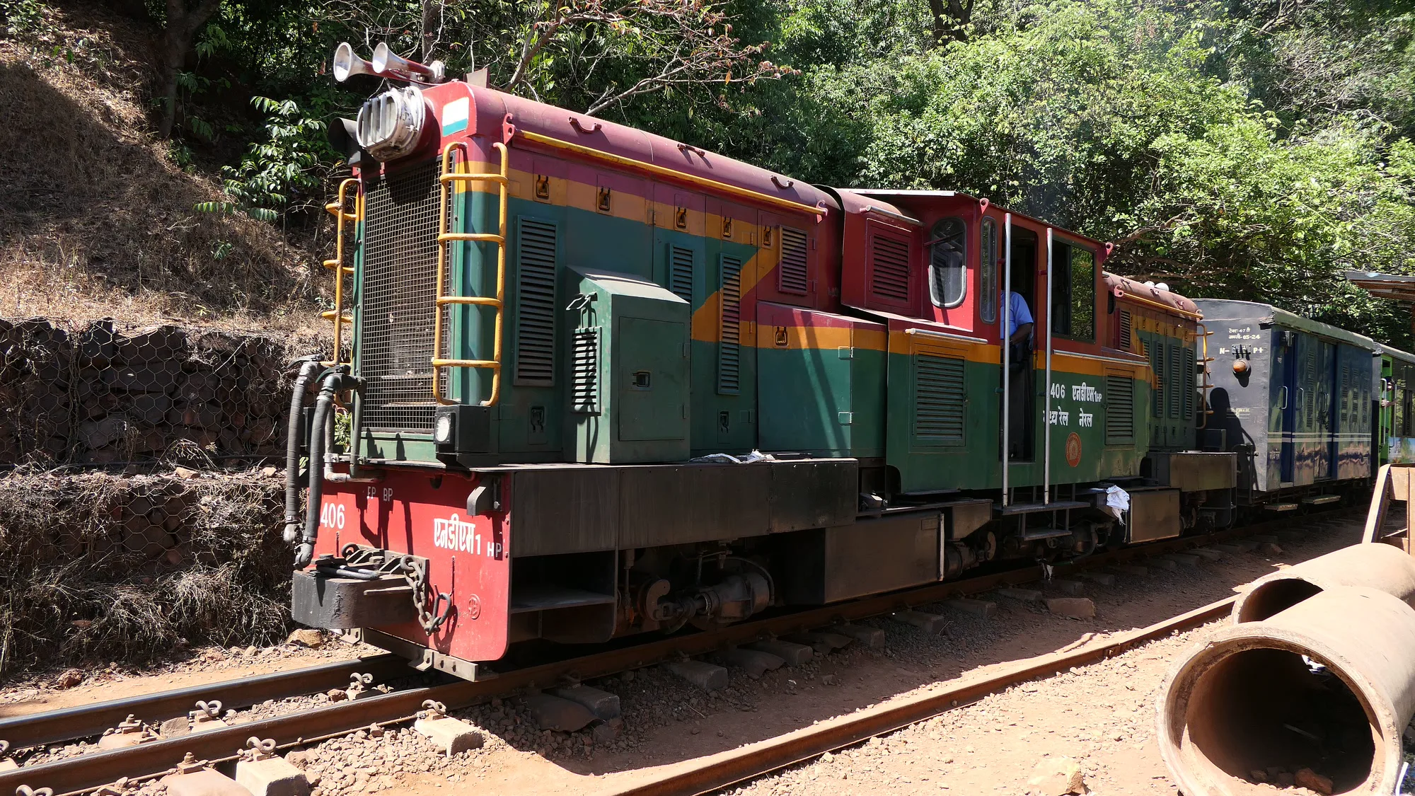 A green and red diesel locomotive of the Matheran toy train pulling blue coaches along a narrow-gauge track through dry forest near Matheran