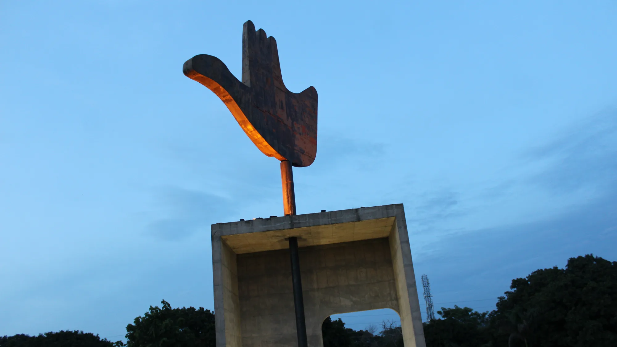 The Open Hand Monument, a large metal hand-shaped wind vane on a concrete frame, against a blue evening sky at the Capitol Complex in Chandigarh
