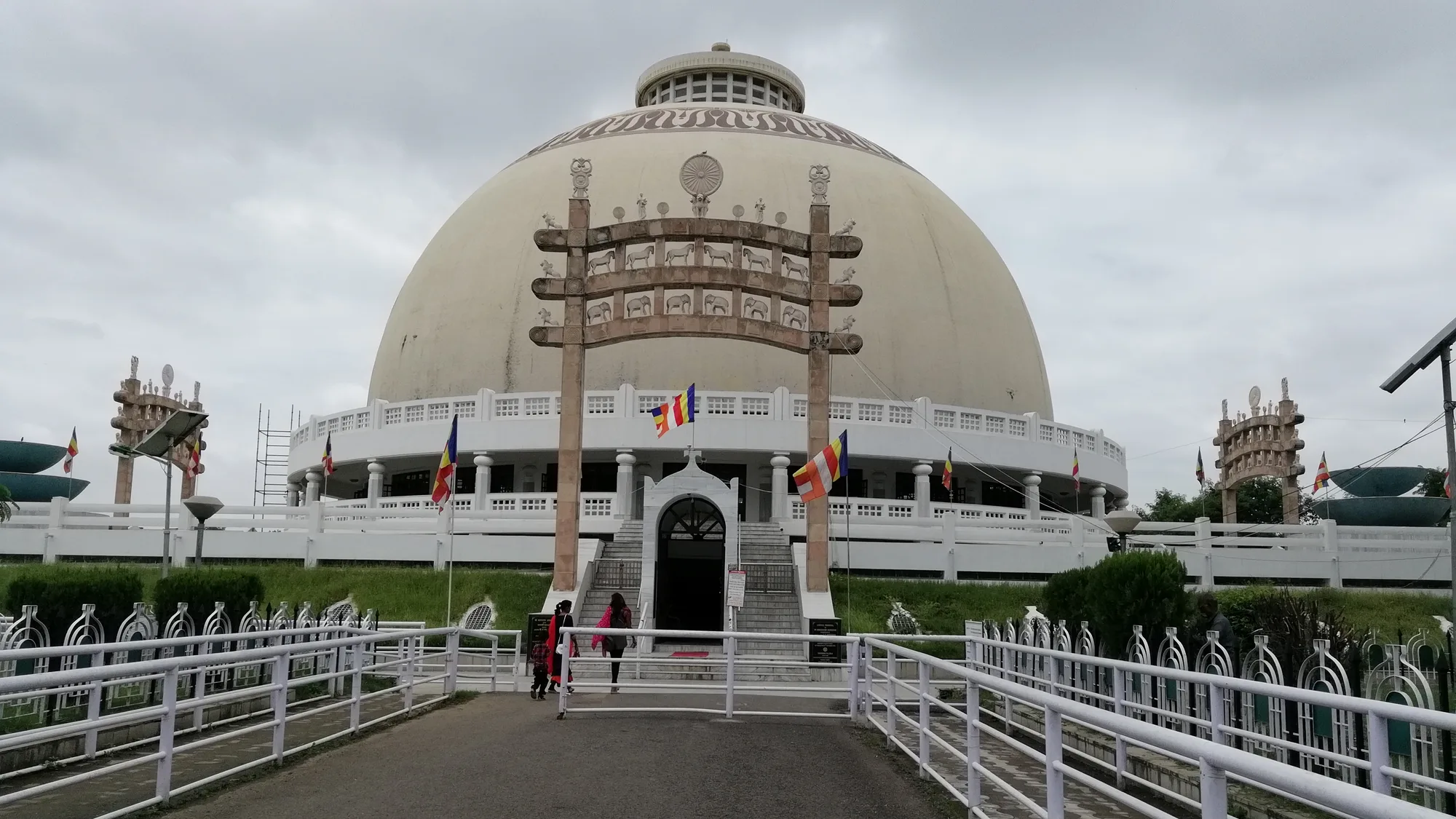 The white hemispherical stupa of Deekshabhoomi in Nagpur with a Sanchi-style stone gateway, Buddhist flags and a railed walkway leading to the entrance