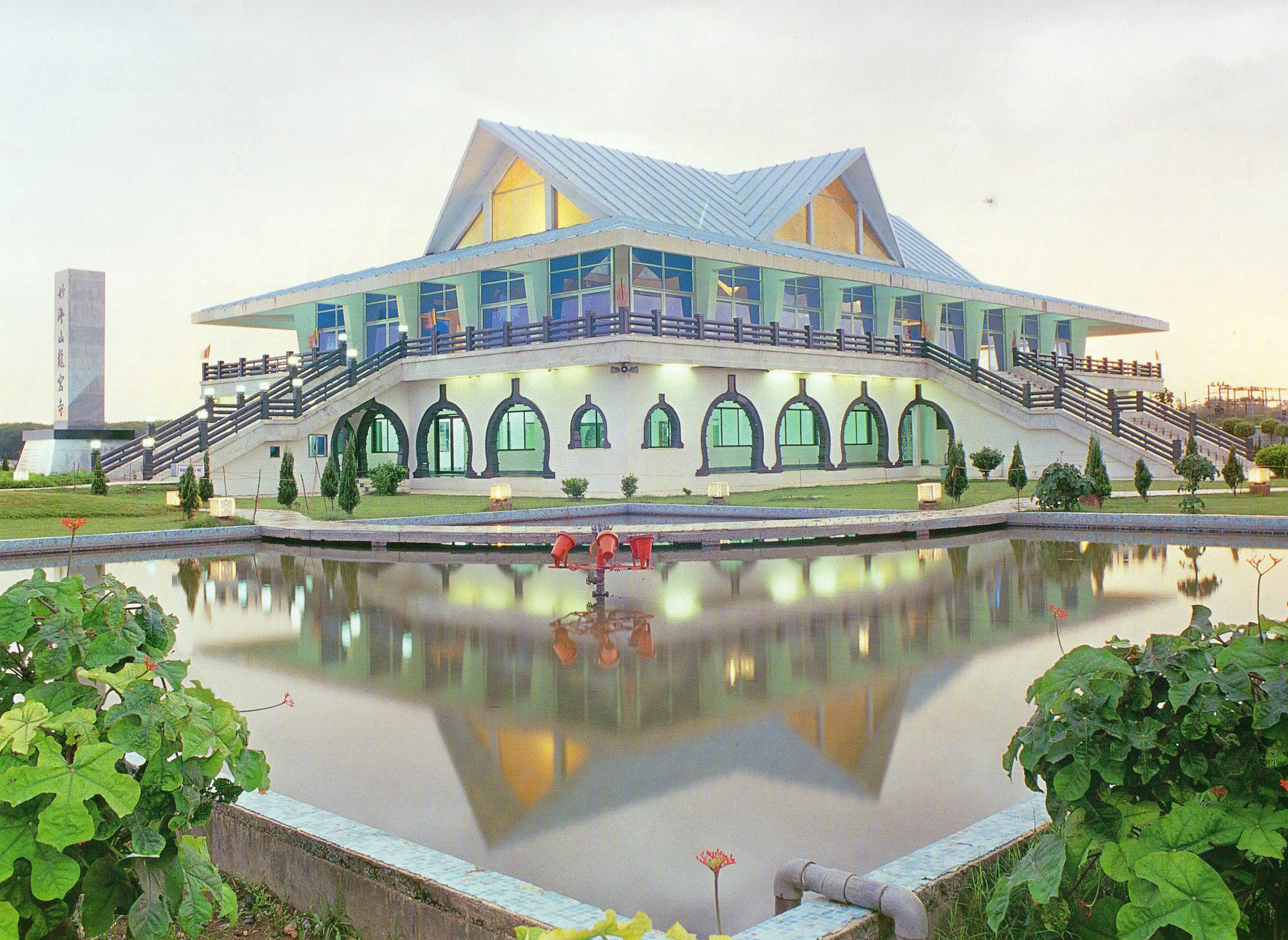 The Dragon Palace Temple at Kamptee near Nagpur, a white and pale-green building with a sloping pagoda-style roof, reflected in a rectangular pool at dusk