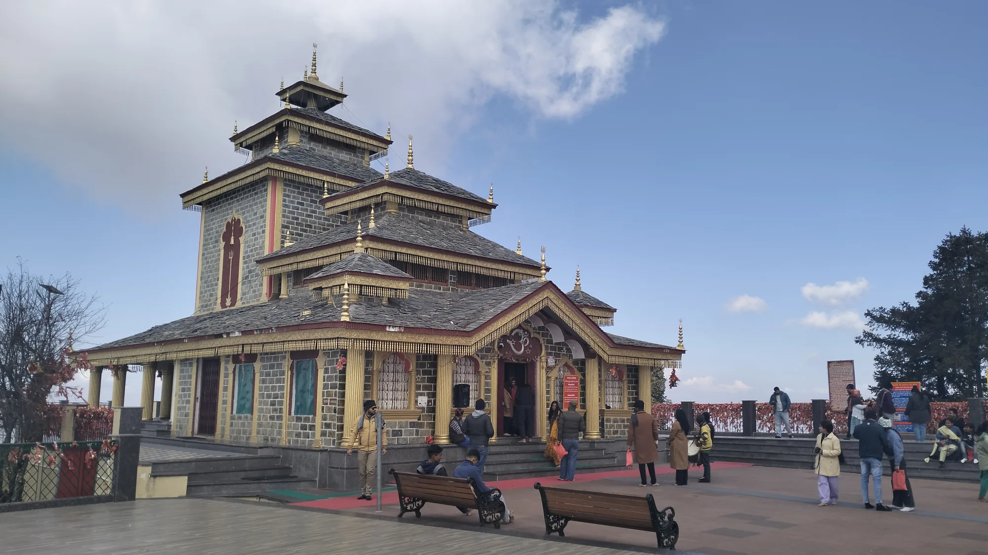 The Surkanda Devi temple near Dhanaulti, a grey stone shrine with tiered slate roofs and gilded finials, with visitors on the paved courtyard under a blue sky