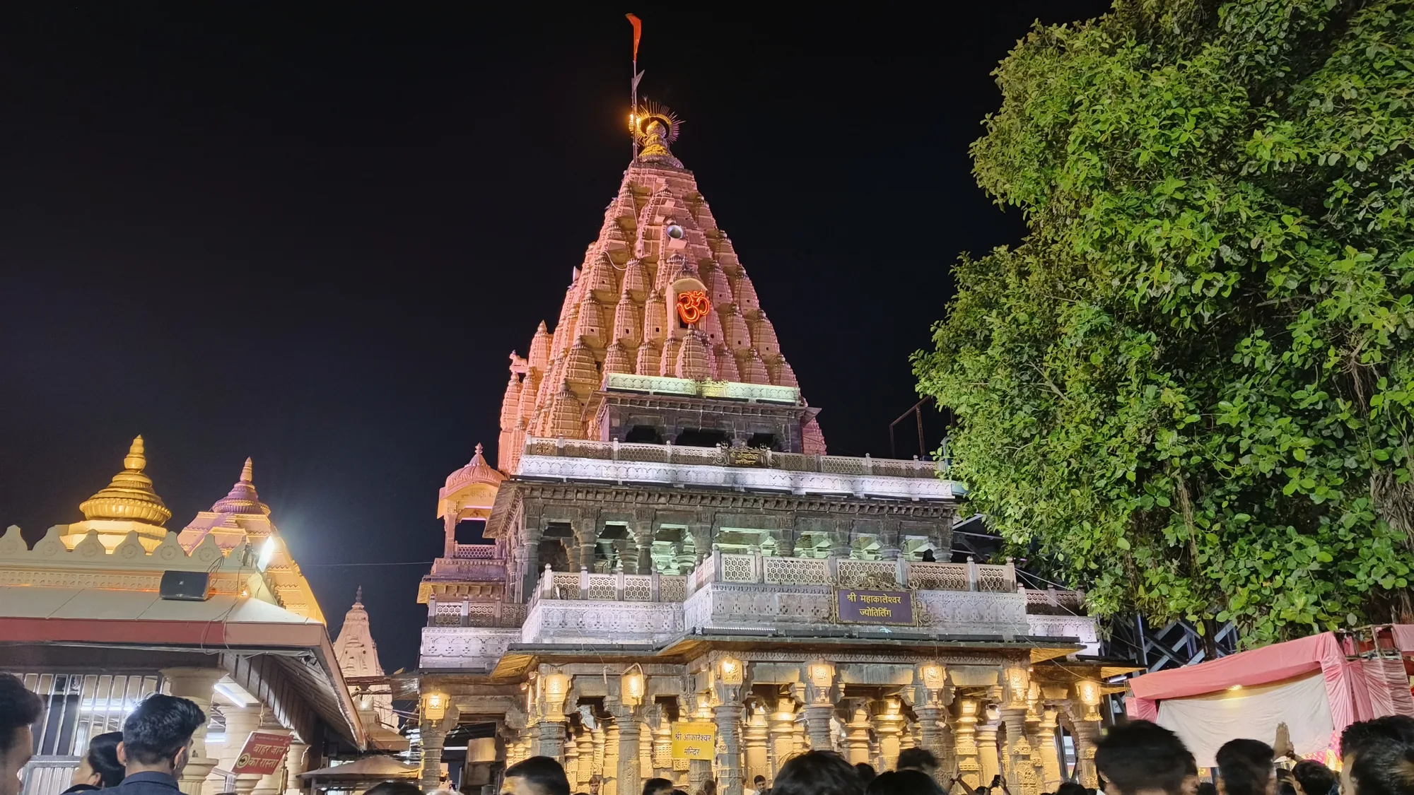 The tall, floodlit spire of the Mahakaleshwar temple in Ujjain at night, with gilded smaller domes on the left, a large tree on the right and devotees in the foreground