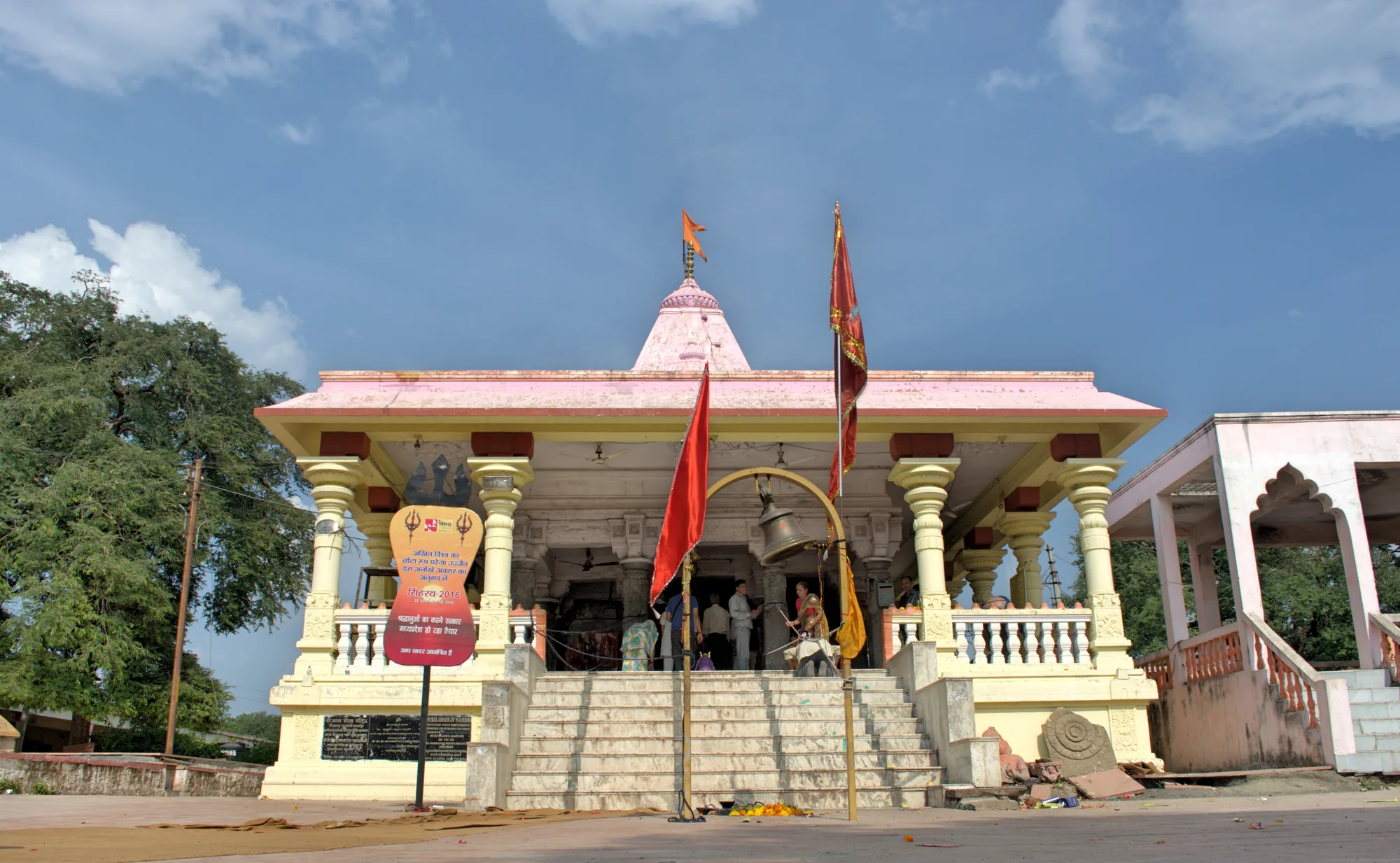 The Kal Bhairav temple in Ujjain, a pale pink and cream shrine with a pillared porch, saffron flags and a wide flight of steps under a blue sky with clouds