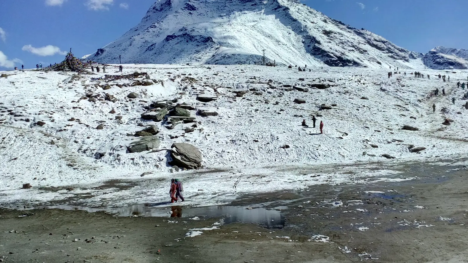 Fresh snow covering the slopes and rocky ground at Rohtang Pass above Manali, with a snow-capped peak behind, visitors walking across the snow and a meltwater pool in front