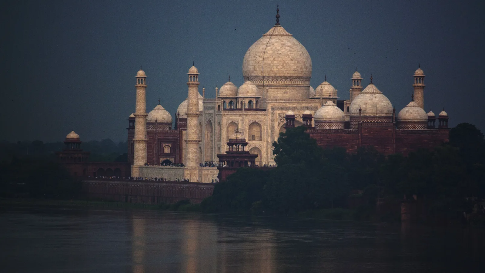 The white marble Taj Mahal and its red sandstone mosque seen from across the Yamuna at dusk, the dome glowing against a dark blue sky above the calm river