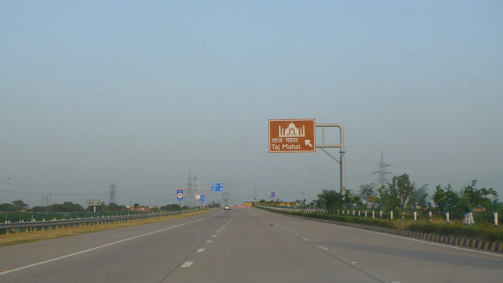 A brown tourist sign for the Taj Mahal over the empty six-lane Yamuna Expressway towards Agra, with flat green fields along the road on a hazy day