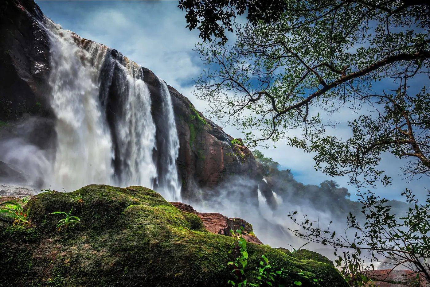 Athirappilly Falls in Kerala seen from below, white water pouring over a dark rock face beside green forest under a blue sky