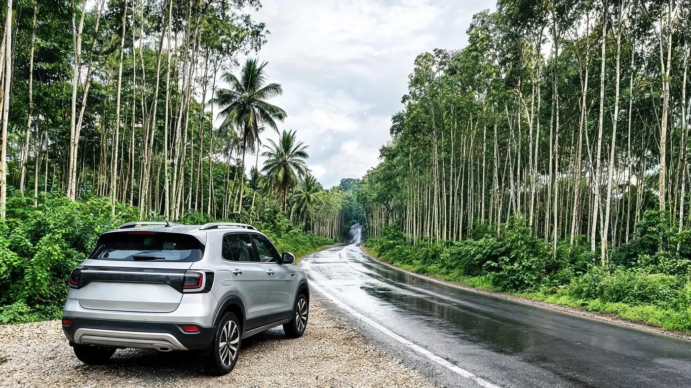 A silver compact SUV parked on the gravel shoulder of a quiet plantation-lined road in central Kerala after rain