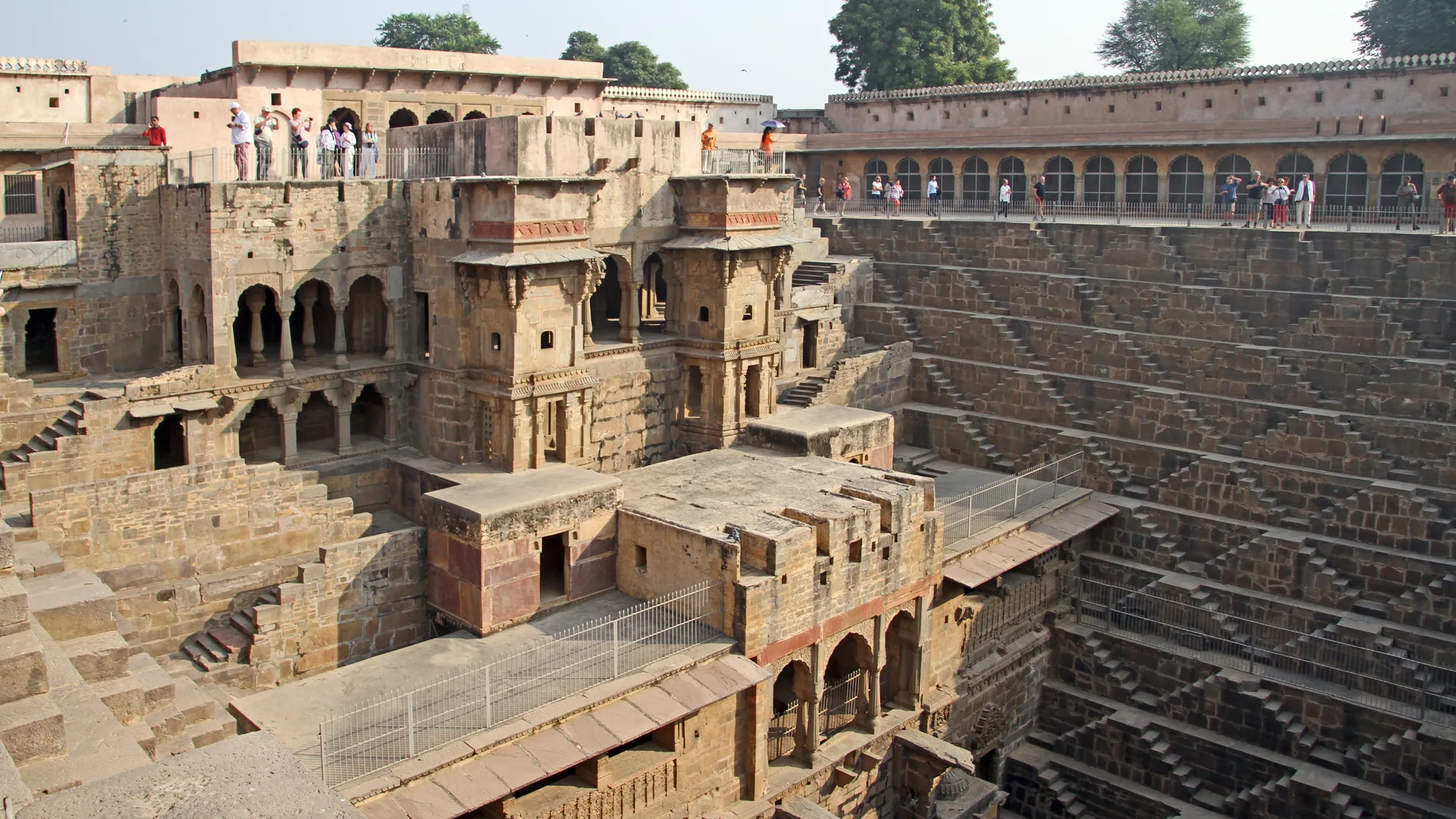 Wide view of Chand Baori stepwell from an upper terrace, with the multi-storey pavilion on one side and visitors along the railings near Jaipur