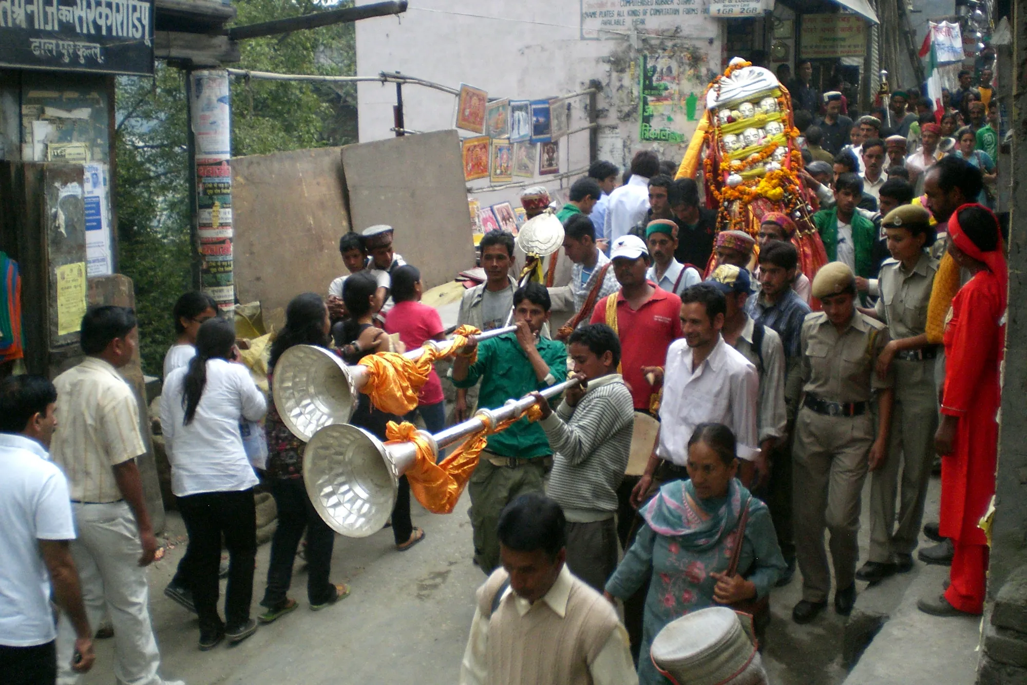 Musicians with brass horns lead a deity palanquin through a crowded Kullu street during Kullu Dussehra