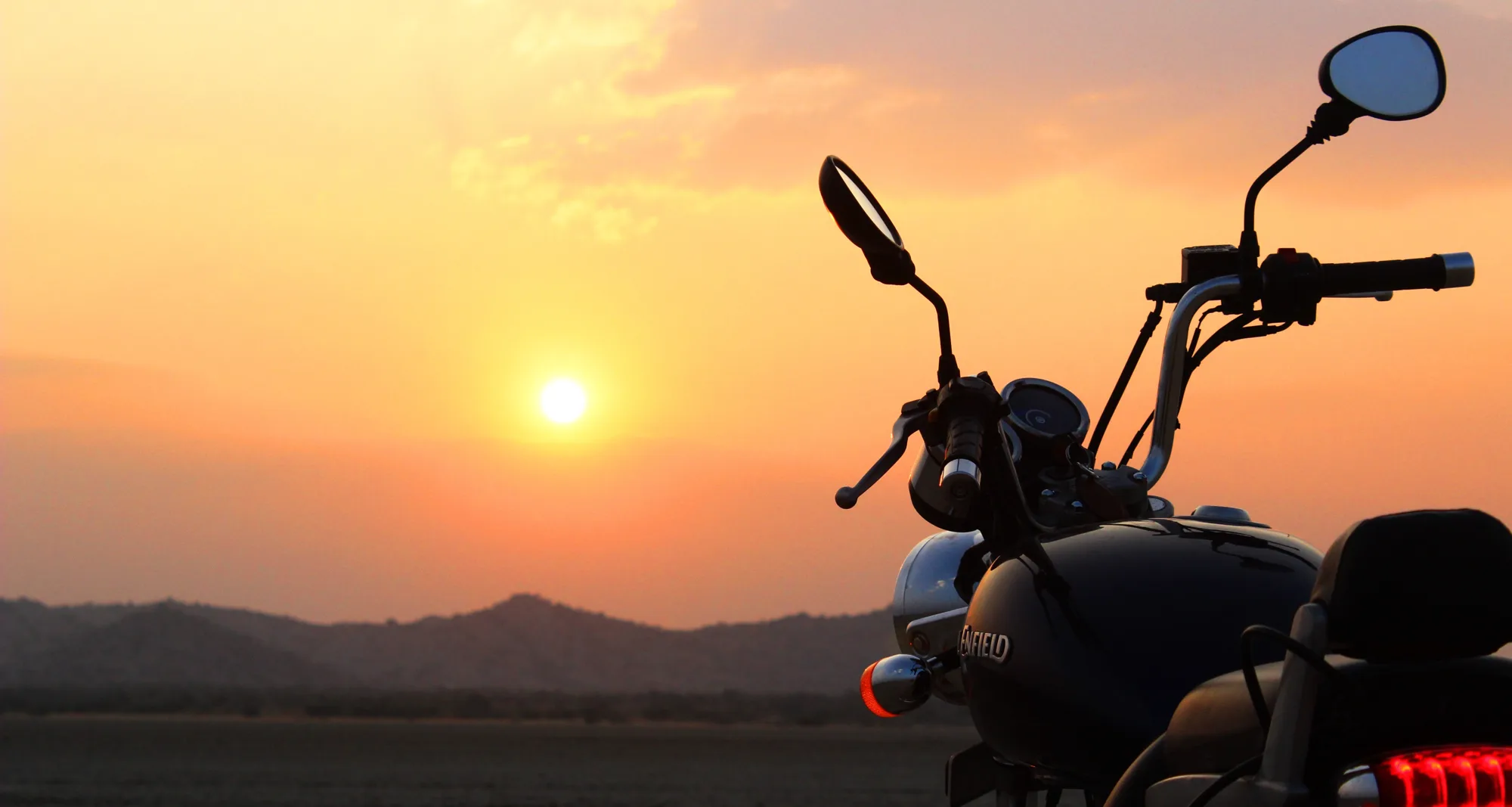 Rear of a motorbike silhouetted against a low orange sun setting over Sambhar Salt Lake and distant hills