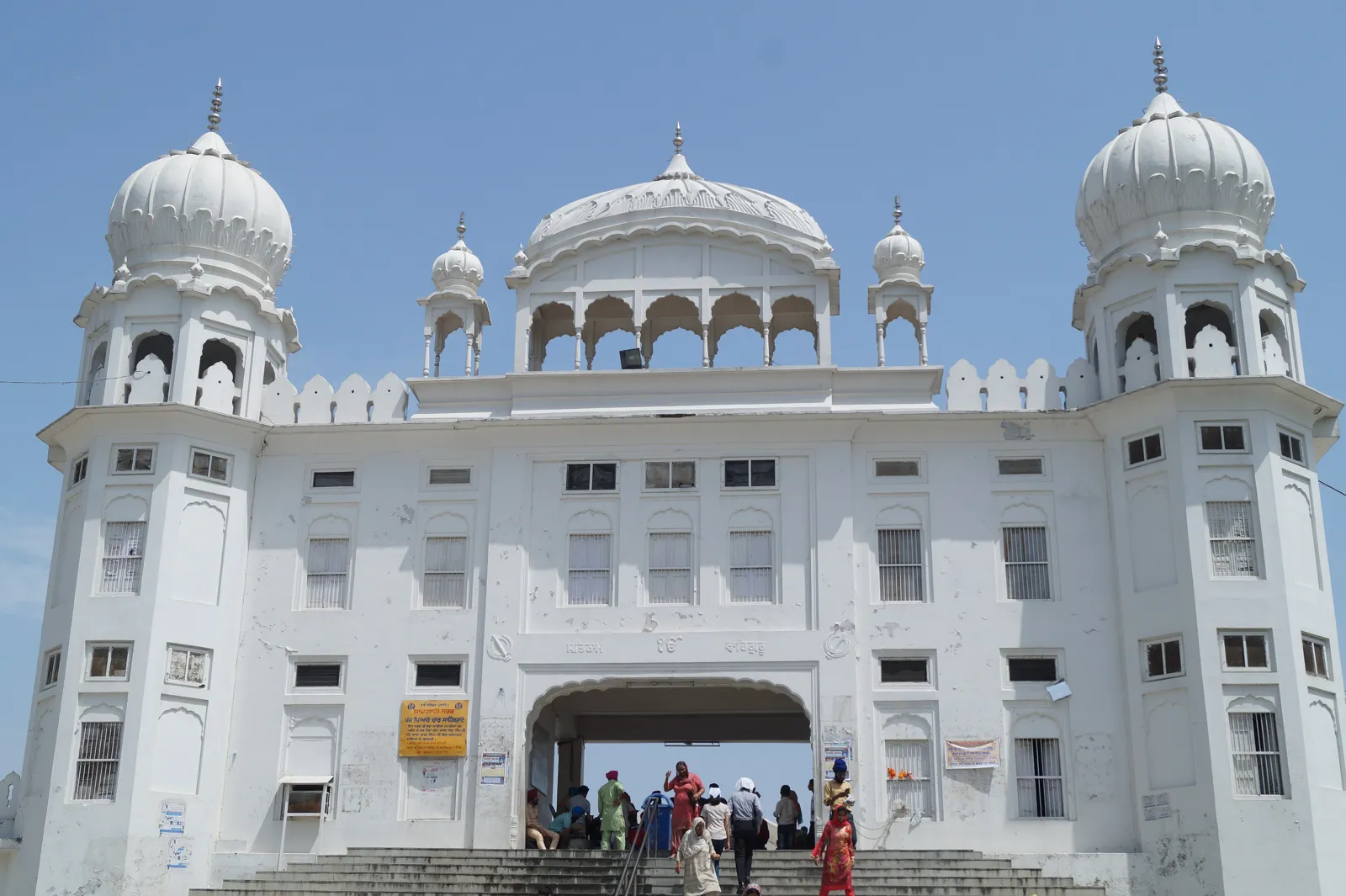 White Takht Sri Kesgarh Sahib building with domes and an arched entrance, with visitors climbing the steps, in Anandpur Sahib