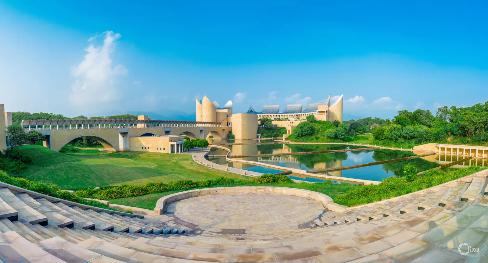 Wide view of the Virasat-e-Khalsa museum complex with sandstone towers, a bridge, a reflecting lake and green lawns under a blue sky