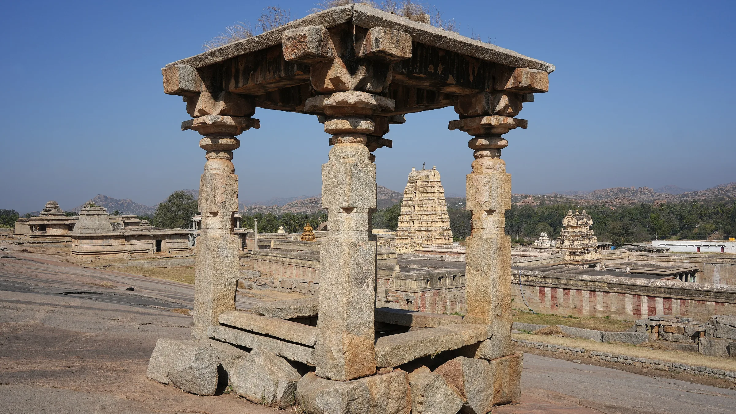 Stone mandapa on Hemakuta Hill with Virupaksha Temple gopuram and Hampi boulder landscape beyond