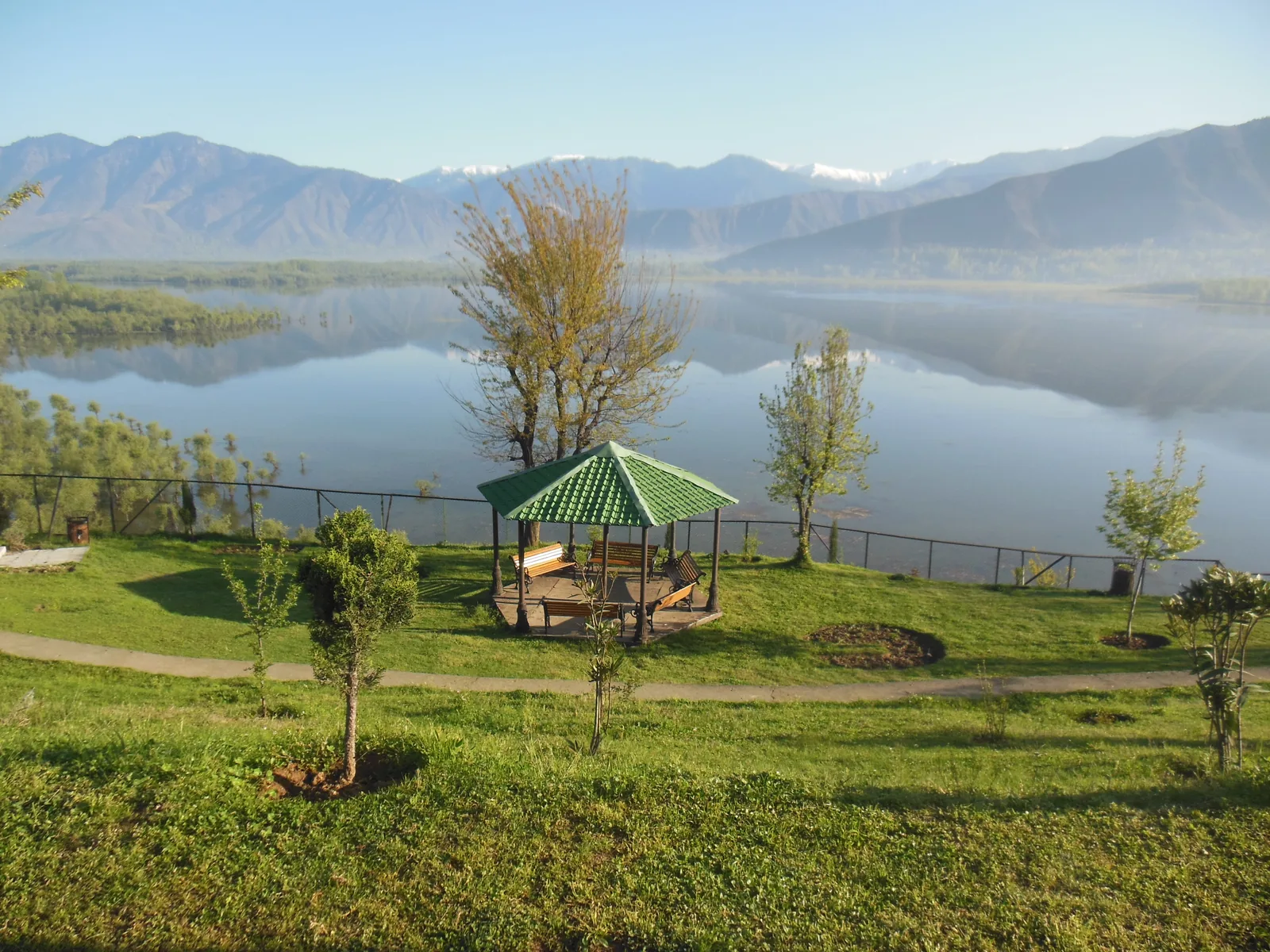Calm water of Wular Lake below a lakeside park, with a green gazebo, young trees and snow-streaked mountains reflected in the lake