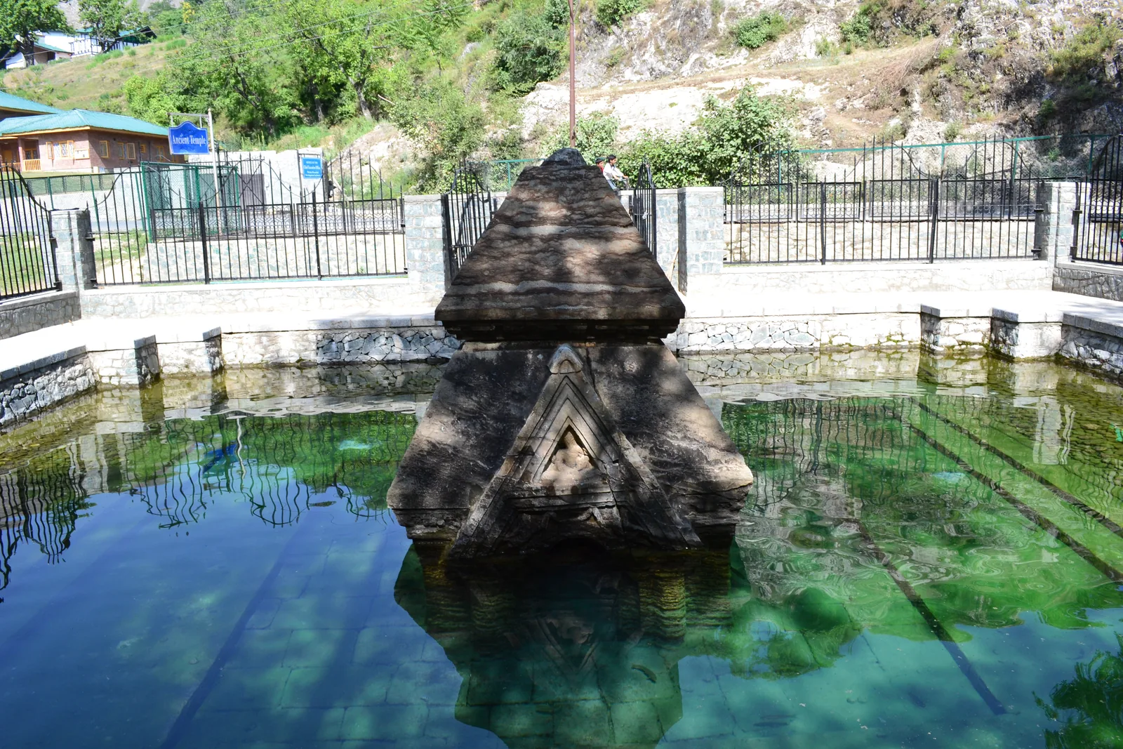 Pyramid-roofed stone temple standing in clear green water in a walled tank near the shore of Manasbal Lake in Ganderbal