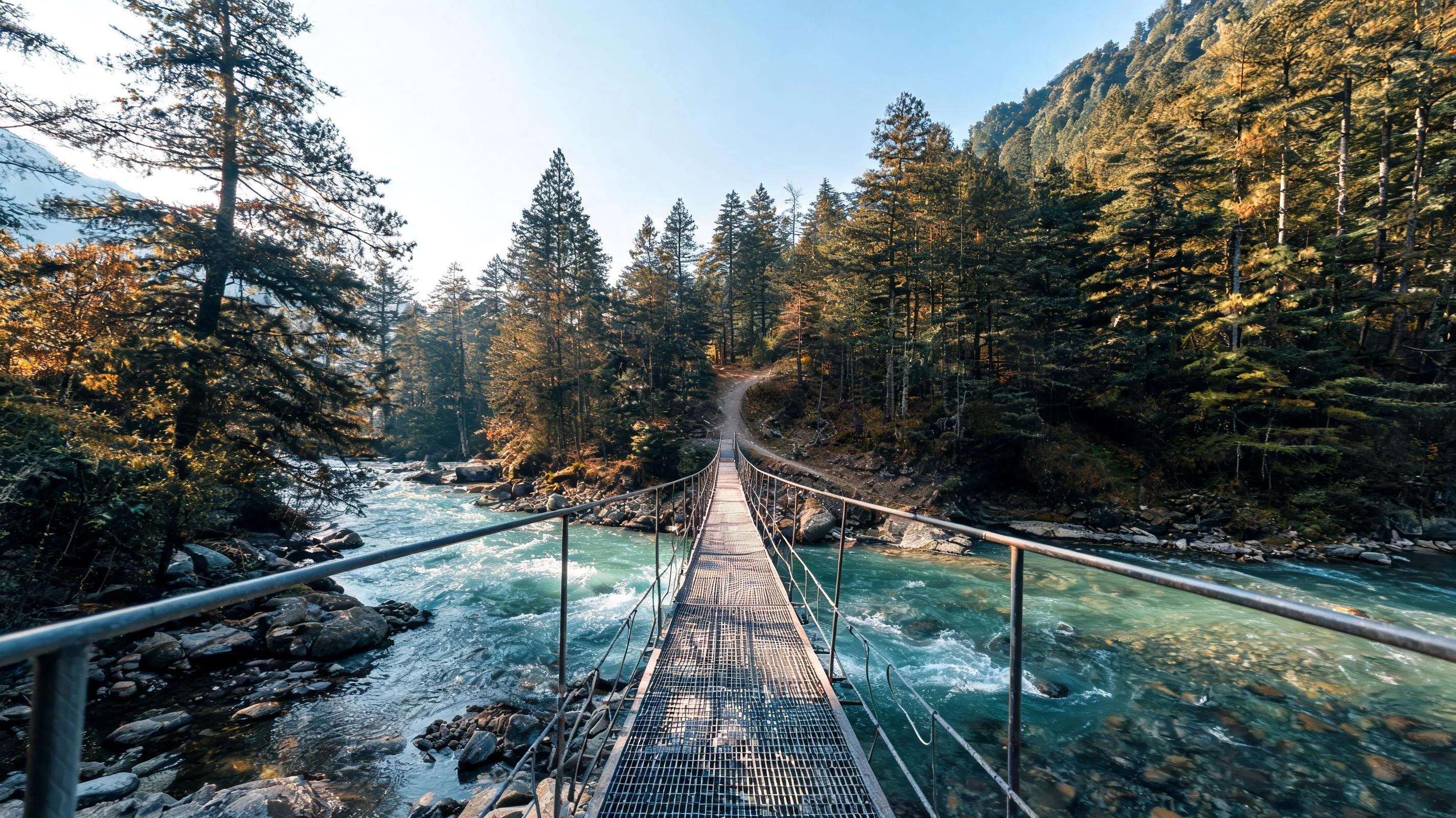 Narrow metal footbridge crossing the turquoise Parvati River toward a pine forest trail opposite Kasol on the way to Chalal