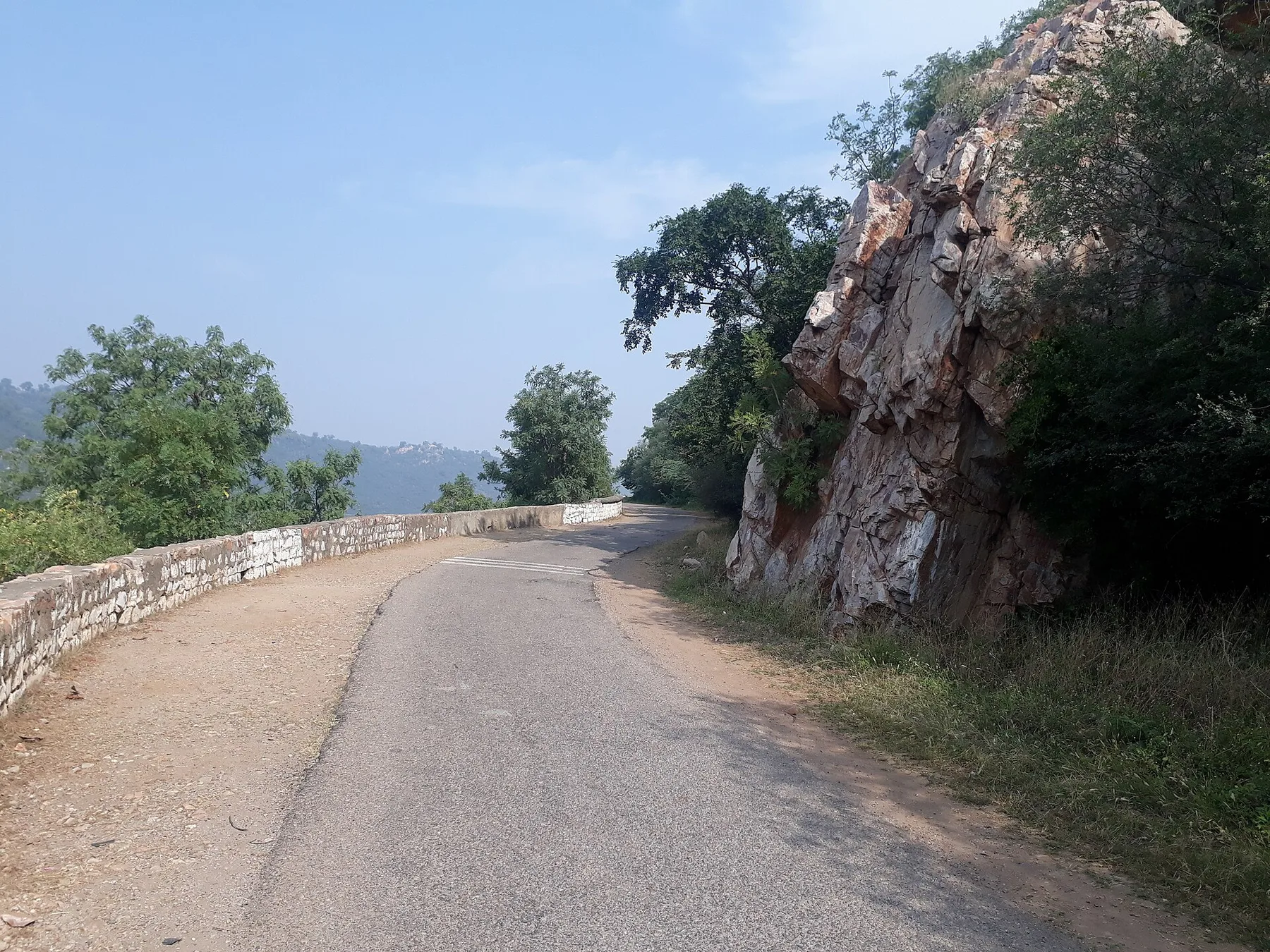 Quiet road with a stone parapet curving past a rocky Aravalli cliff in the forest near Alwar, on the drive from Gurugram to Sariska