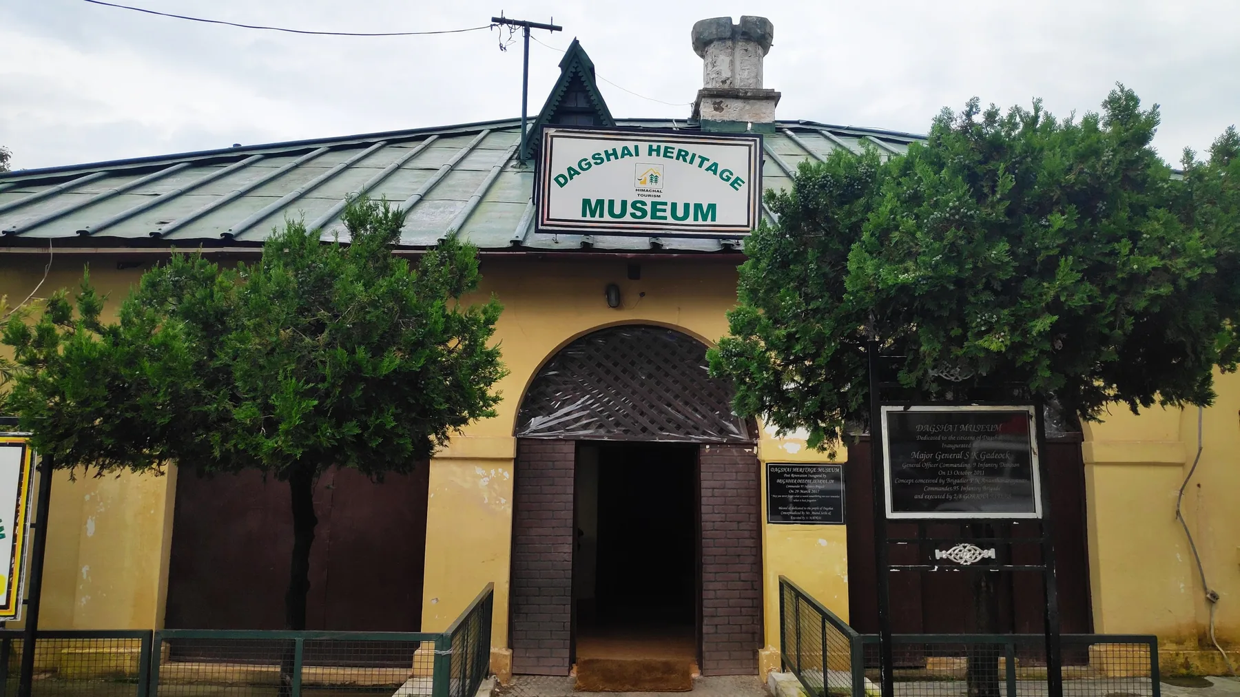 Yellow front of the Dagshai Heritage Museum with an arched wooden door, green roof and trees on either side