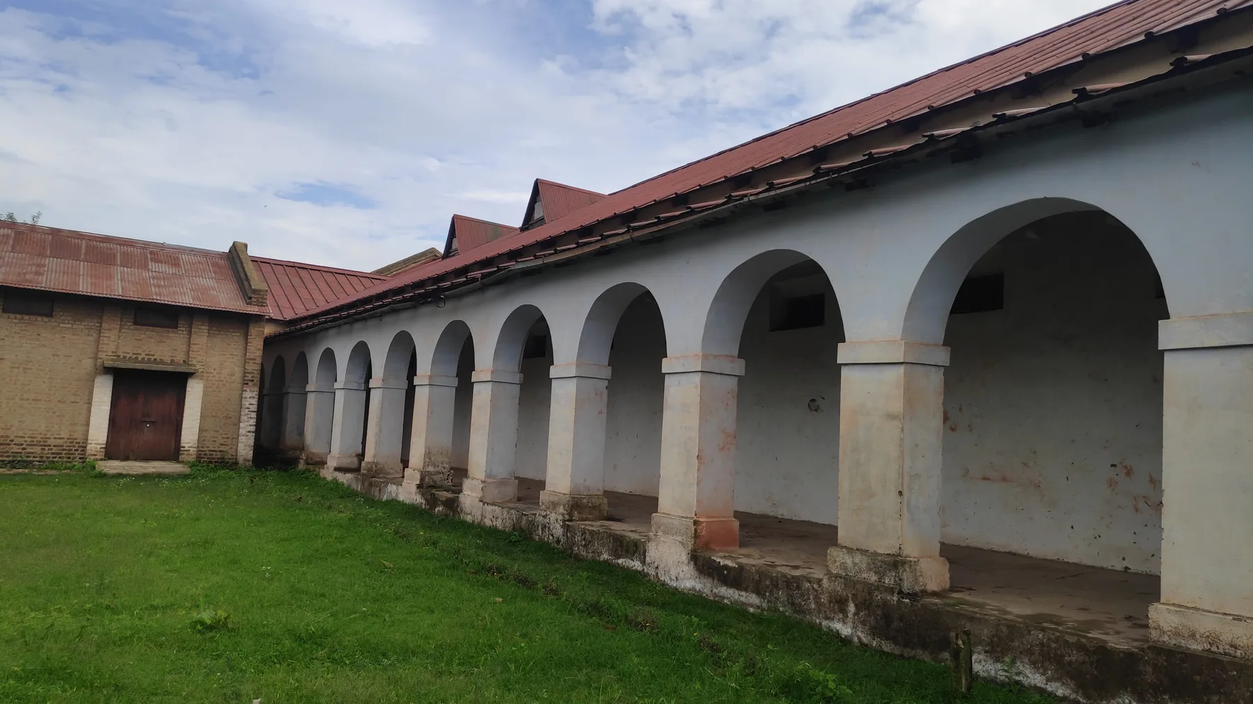 Whitewashed arched verandah of the old jail buildings at Dagshai beside a green lawn under a cloudy sky