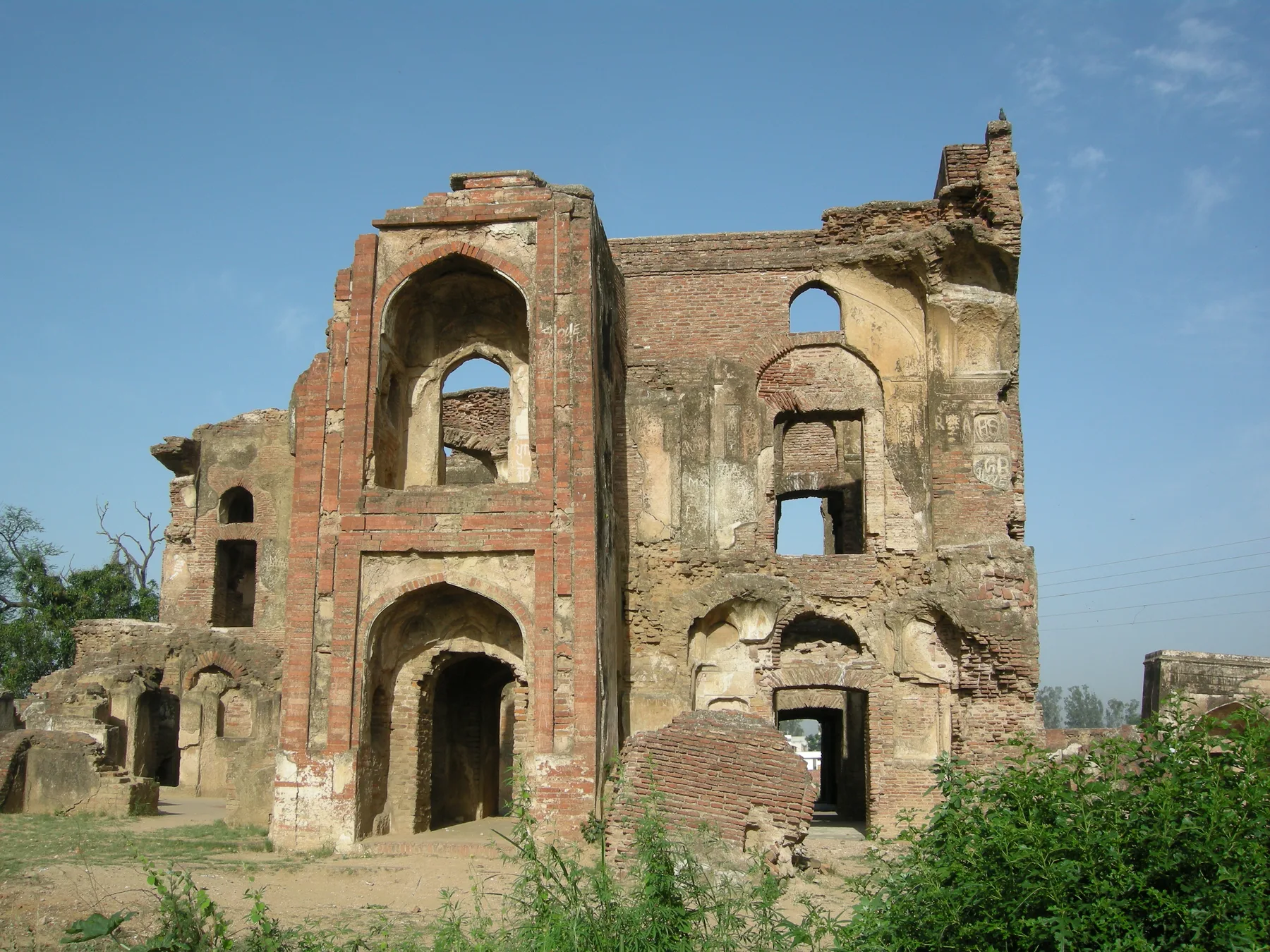 Roofless red-brick ruins of a Mughal palace building at Aam Khas Bagh in Sirhind, with arched openings and green shrubs in front