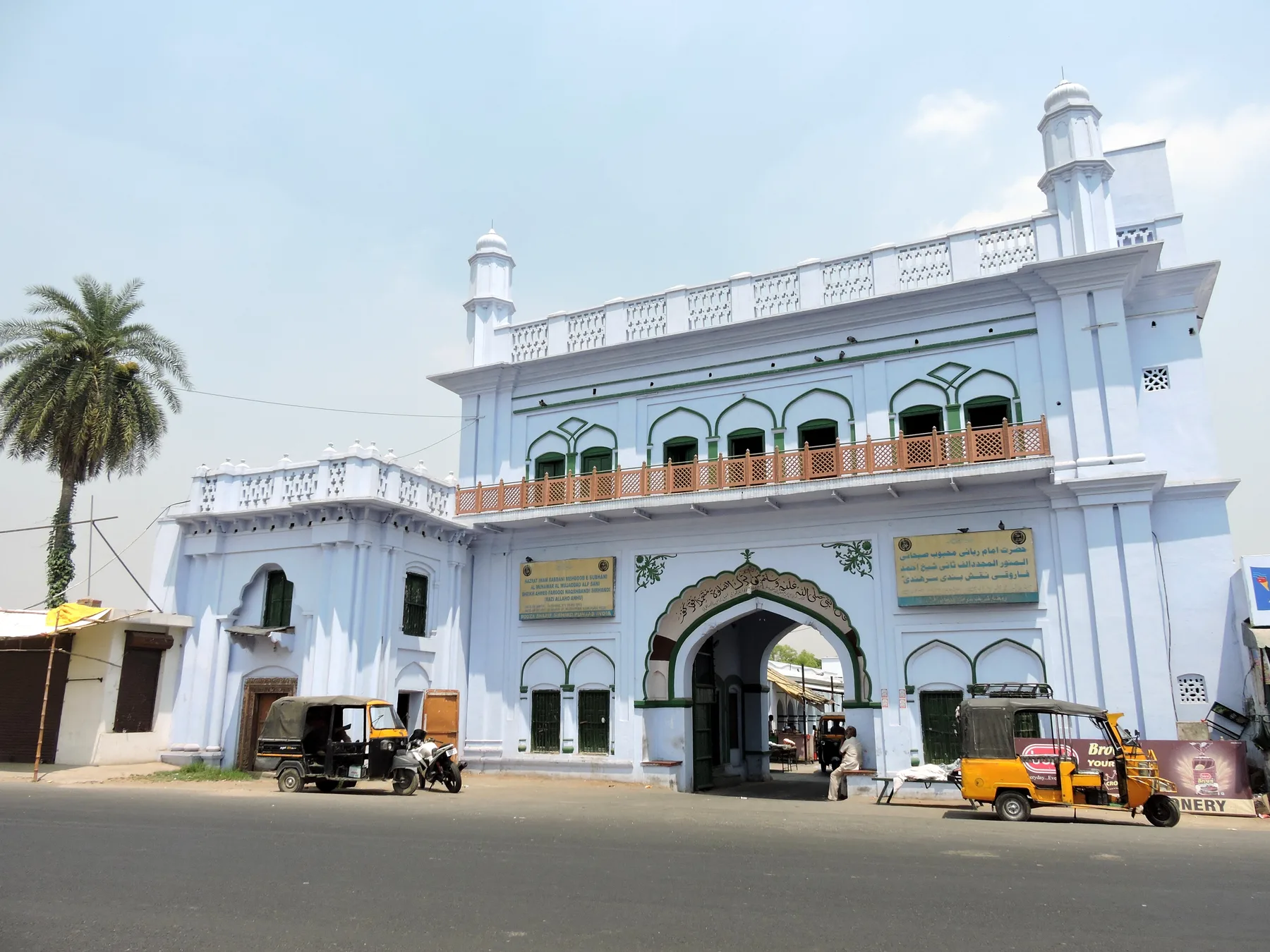The white arched entrance gateway of Rauza Sharif in Sirhind, with small domed turrets on top and a palm tree beside it