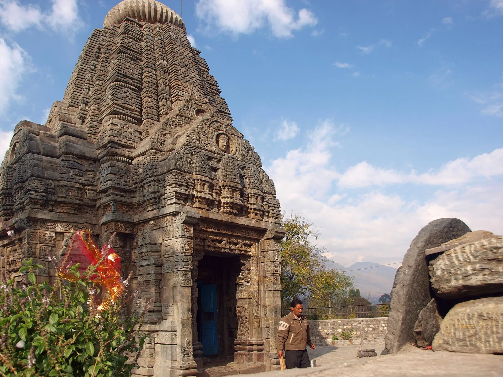 The carved stone shikhara of the Basheshar Mahadev temple at Bajaura, with an amalaka on top, a blue doorway and hills in the background