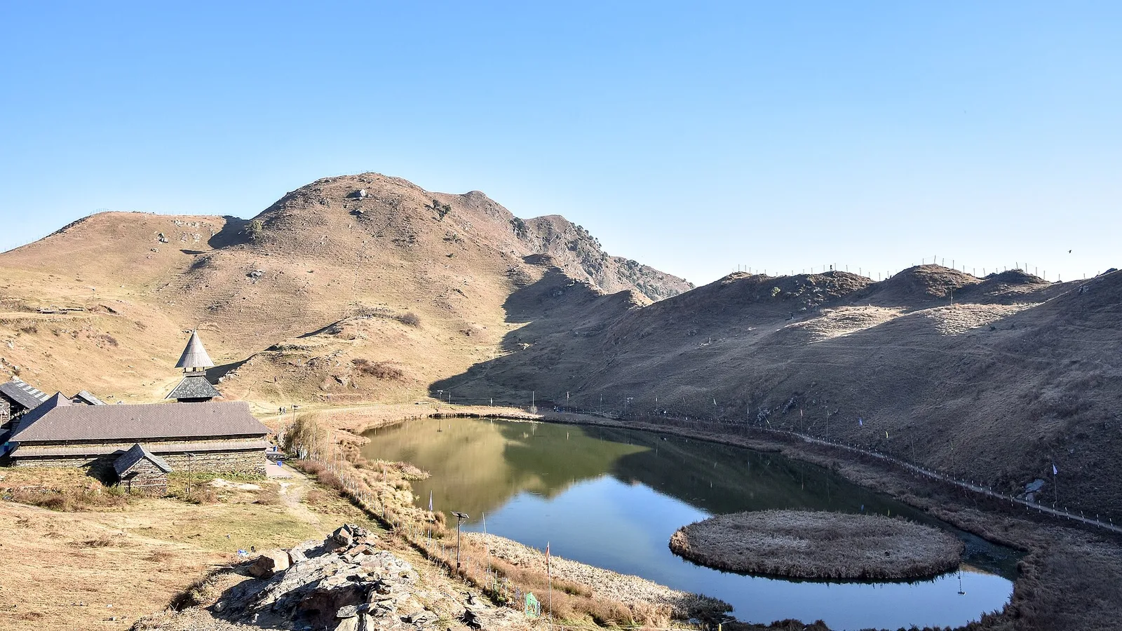 Prashar Lake in late autumn with its round floating island, the pagoda temple on the left and golden-brown grassy ridges under a clear sky, a ride from a bike rental in Bhuntar
