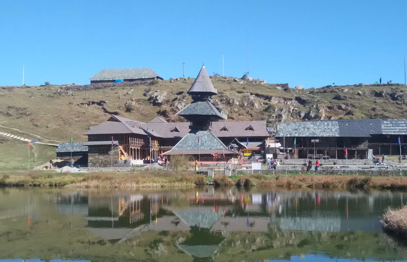 The three-tiered wooden pagoda temple of Rishi Prashar reflected in the still lake water, with slate roofs and a dry grassy hillside behind