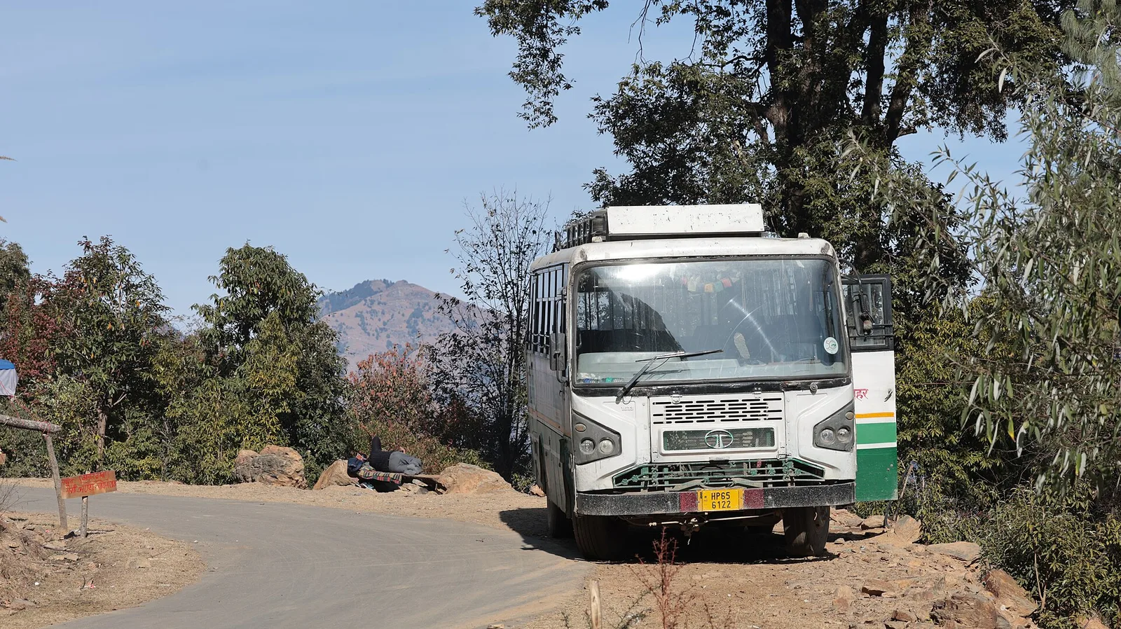 A paved mountain road bending past oak trees near Parashar, with a parked HRTC bus and a brown hilltop in the distance