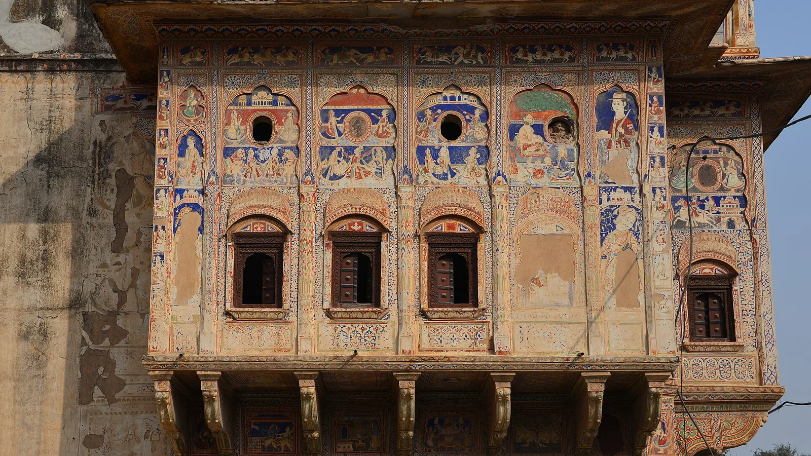 The painted upper facade of an old haveli in Mandawa, Shekhawati, with fresco panels of figures and flowers, arched windows and carved brackets, a day trip by self-drive car rental in Jaipur