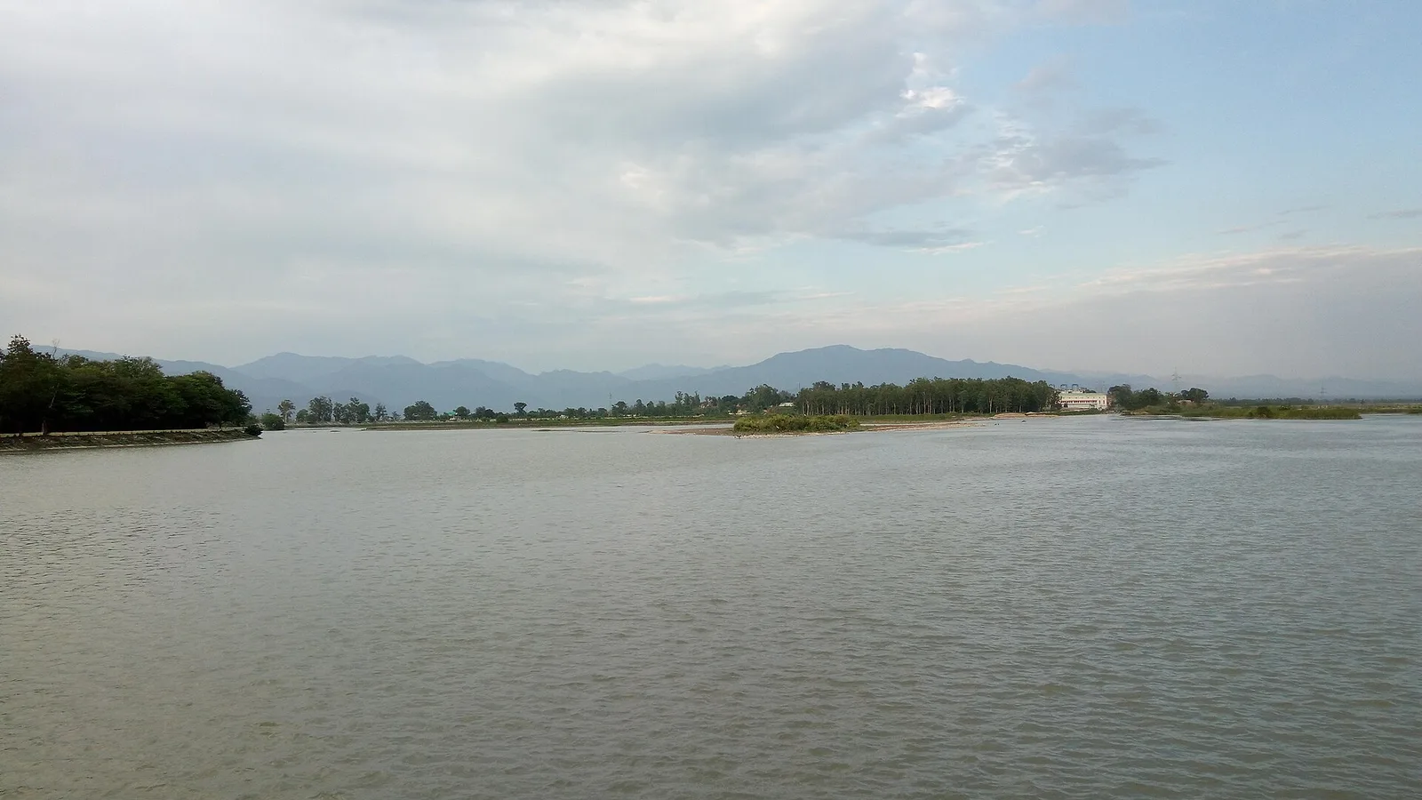 The wide reservoir behind Asan Barrage under a cloudy sky, with low green islands and the Shivalik hills on the horizon