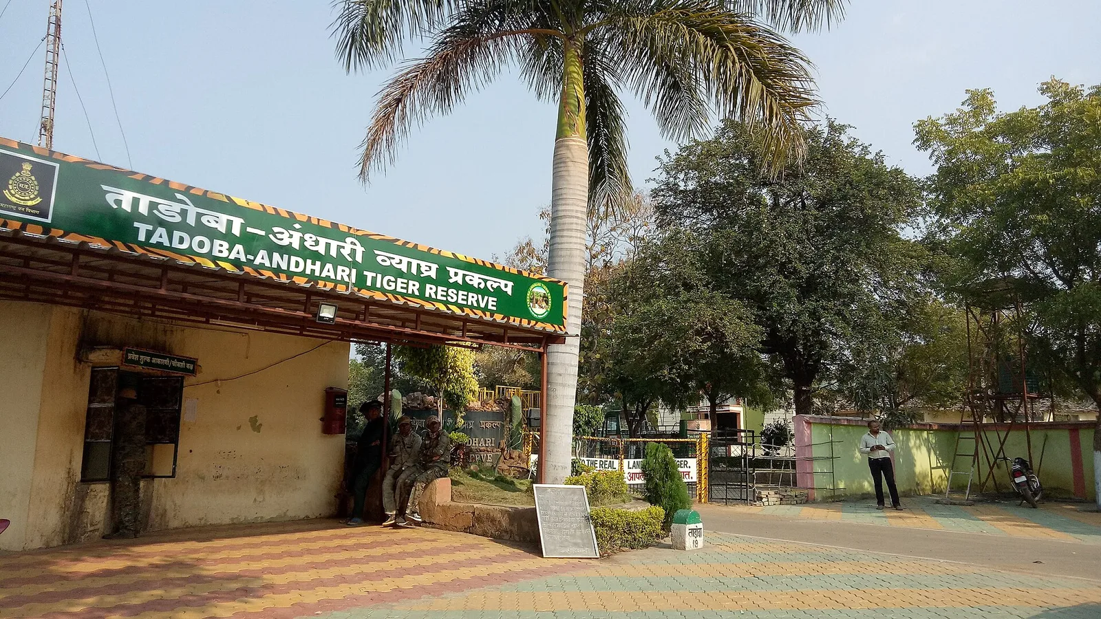The reception building at Moharli Gate of Tadoba Andhari Tiger Reserve, with a green signboard, palm trees and a paved forecourt