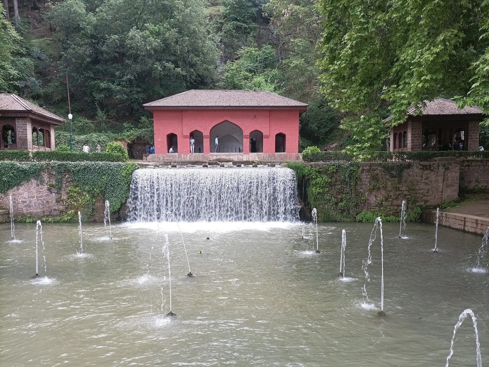 A cascade falling from a pink pavilion into a fountain pool at the Achabal Mughal garden, with forested hills behind