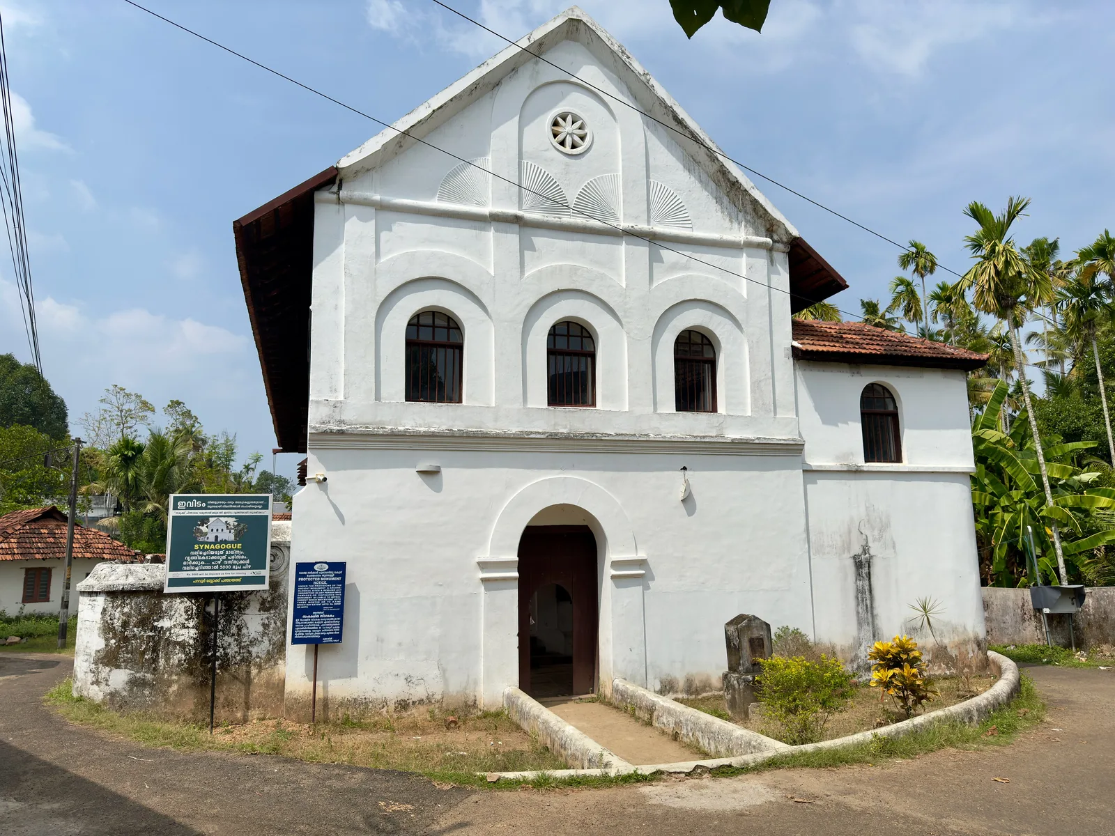 The whitewashed gabled facade of the Chendamangalam Synagogue with arched windows, a small garden and coconut palms