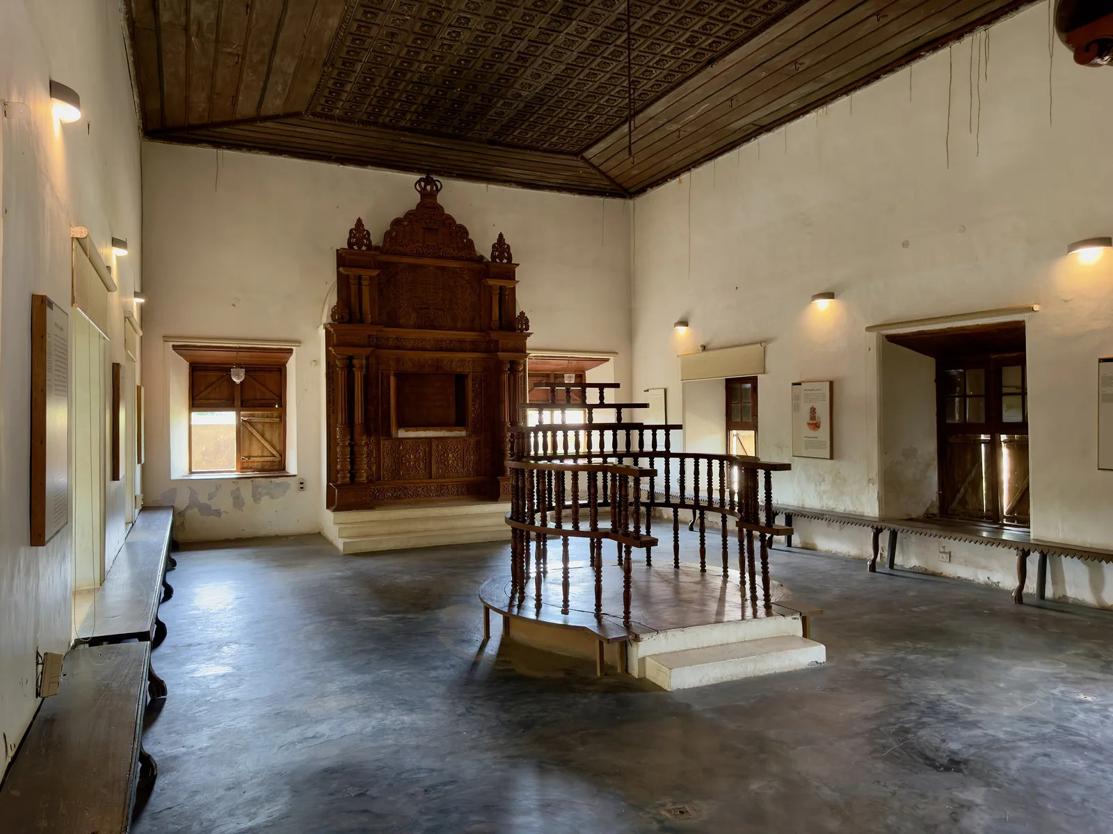 Inside the Paravur Synagogue museum: a carved teak ark against a whitewashed wall, a railed reading platform and a dark wooden ceiling
