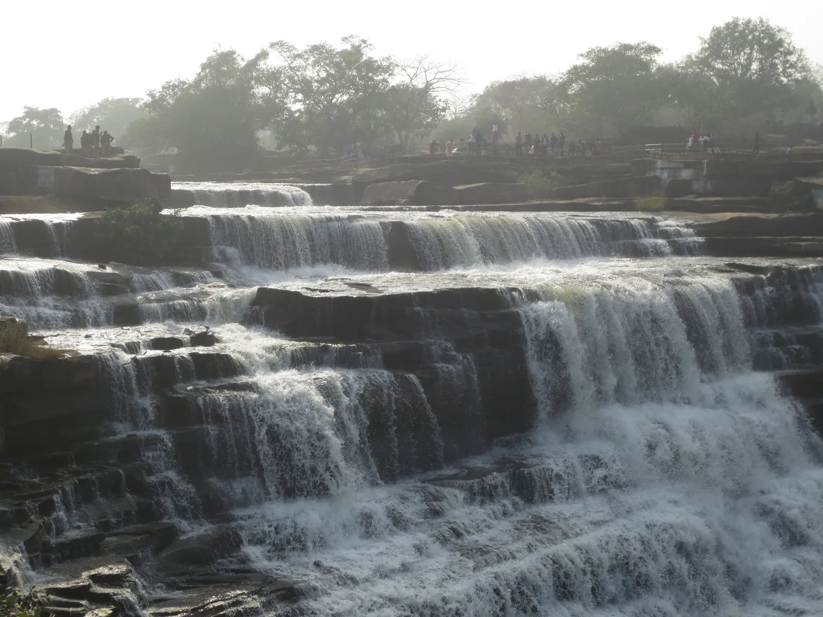 Rajdari waterfall spilling over wide, stepped sandstone ledges, with visitors standing along the railings at the top