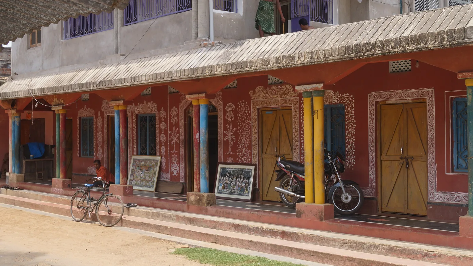 A row of Raghurajpur village houses with red walls covered in white murals, colourful pillars and framed Pattachitra paintings on the veranda, a day trip by self-drive car rental in Bhubaneswar