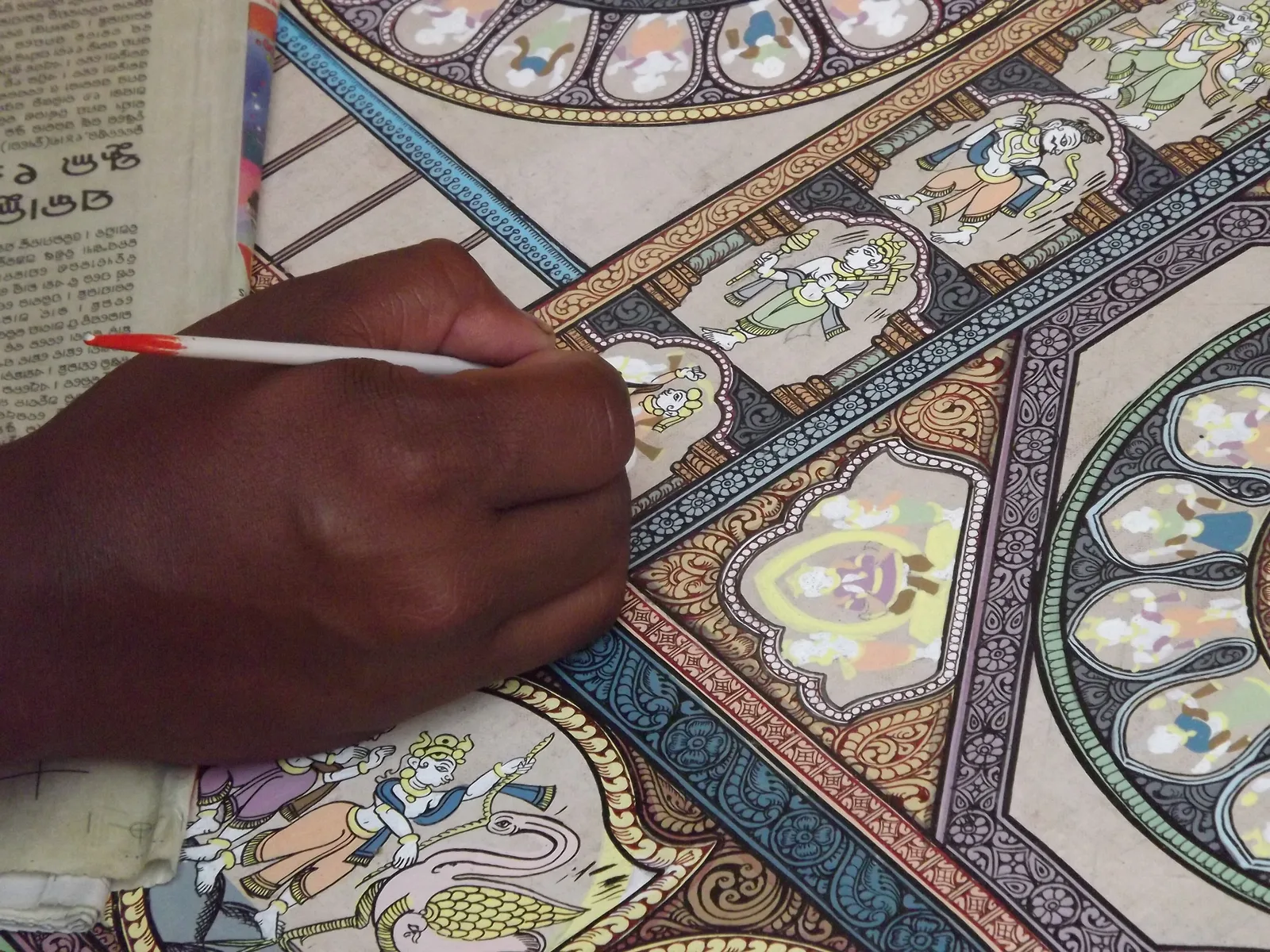 Close-up of an artist's hand painting fine details on a Pattachitra with a thin brush in Raghurajpur