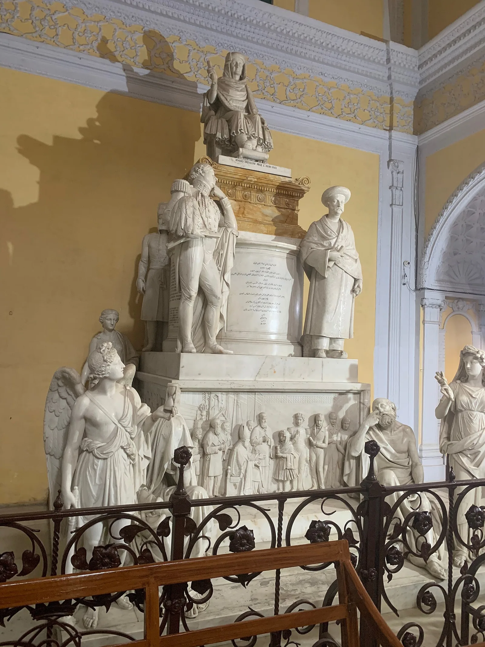 The white marble monument over Begum Samru's tomb inside the Sardhana basilica, with her seated figure at the top and carved figures and a relief panel below, behind an iron railing