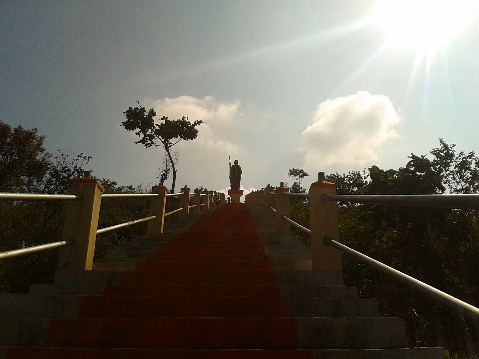 Steep red-carpeted steps with metal railings climbing towards a statue on top of Kurisumala hill at Vagamon, with the sun behind