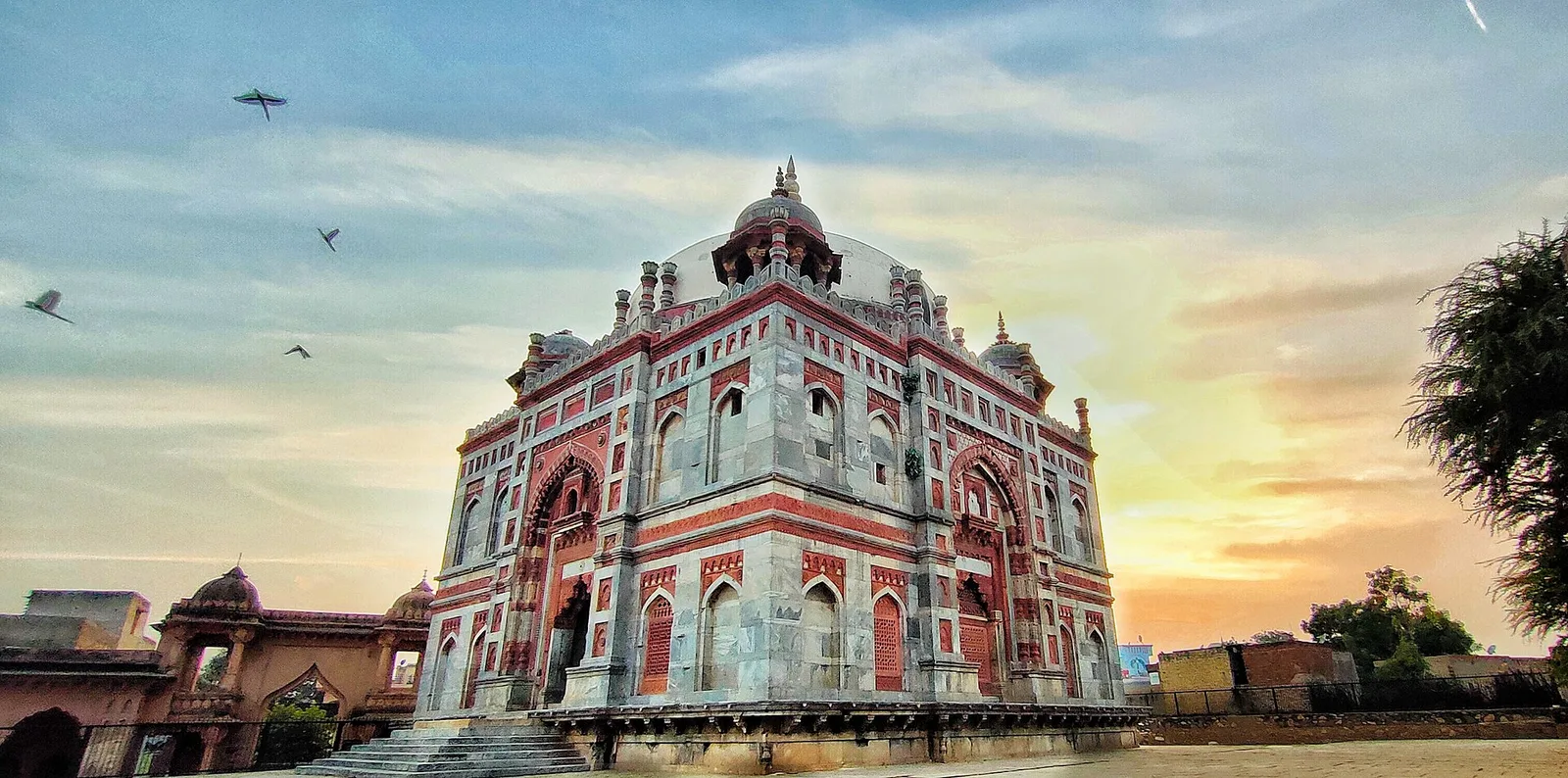 The square tomb of Ibrahim Khan Sur in Narnaul, built in red and grey stone with a white dome, small corner cupolas and birds in an evening sky