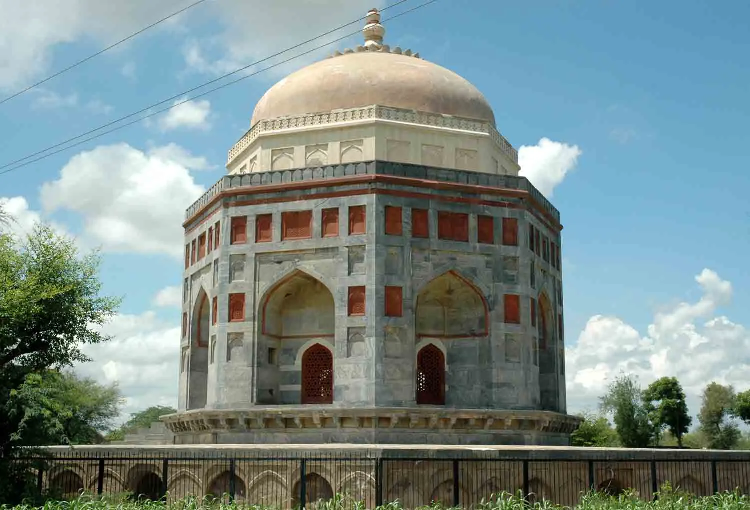The octagonal tomb of Shah Quli Khan in Narnaul, in grey stone with red sandstone panels and a pale dome, seen behind a low railing