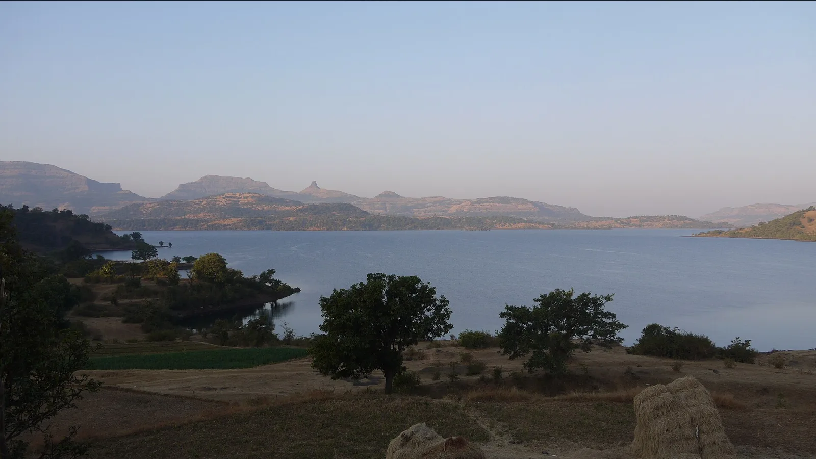 Arthur Lake at Bhandardara on a clear morning, with flat-topped Sahyadri hills behind the water and trees on a dry grassy bank, a trip by self-drive car rental in Mumbai