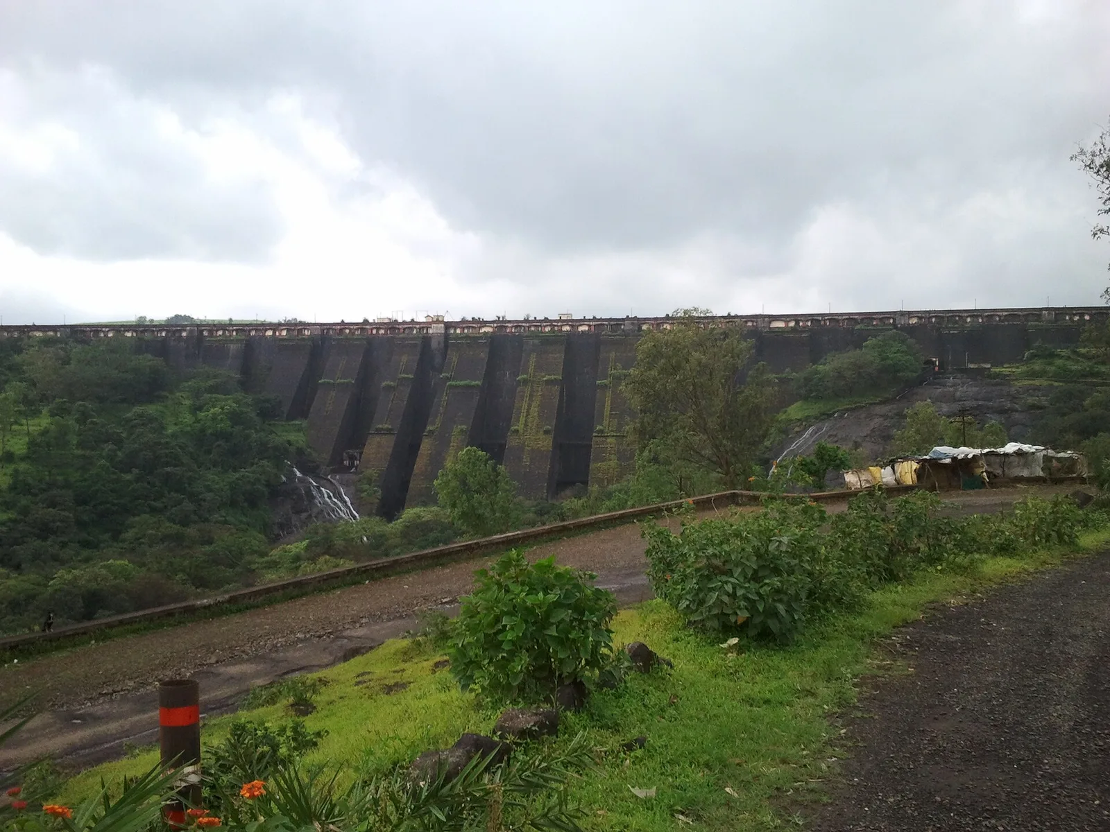 The long grey masonry wall of the Wilson Dam at Bhandardara under monsoon clouds, with green slopes and a road along the bank
