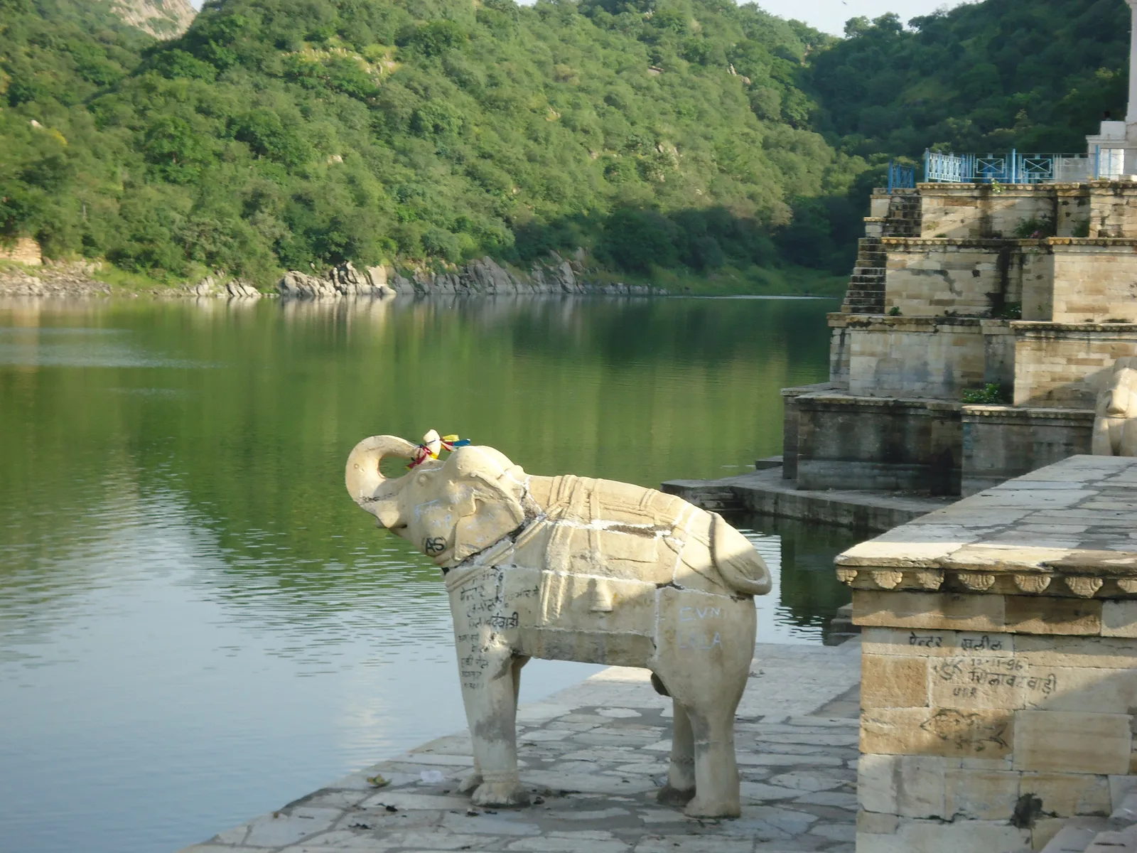A white marble elephant statue on the Jaisamand dam, standing on a stone platform beside the lake with green hills behind