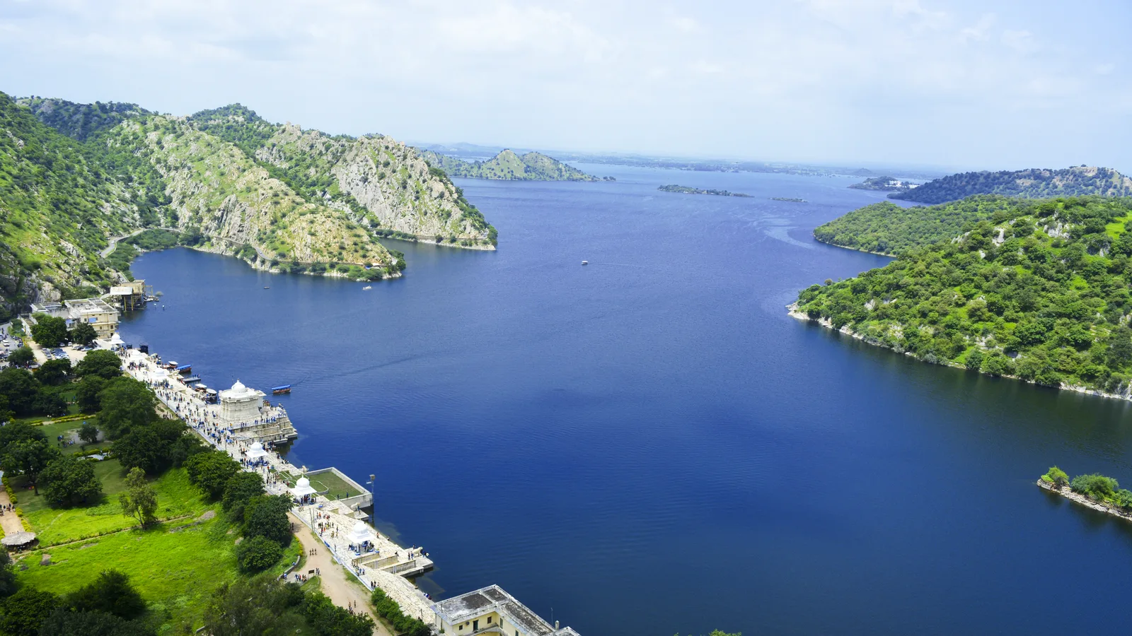 Aerial view of Jaisamand Lake near Udaipur, a long blue lake between green Aravalli hills with the white marble dam and its pavilions along the left shore, a trip by self-drive car rental in Udaipur